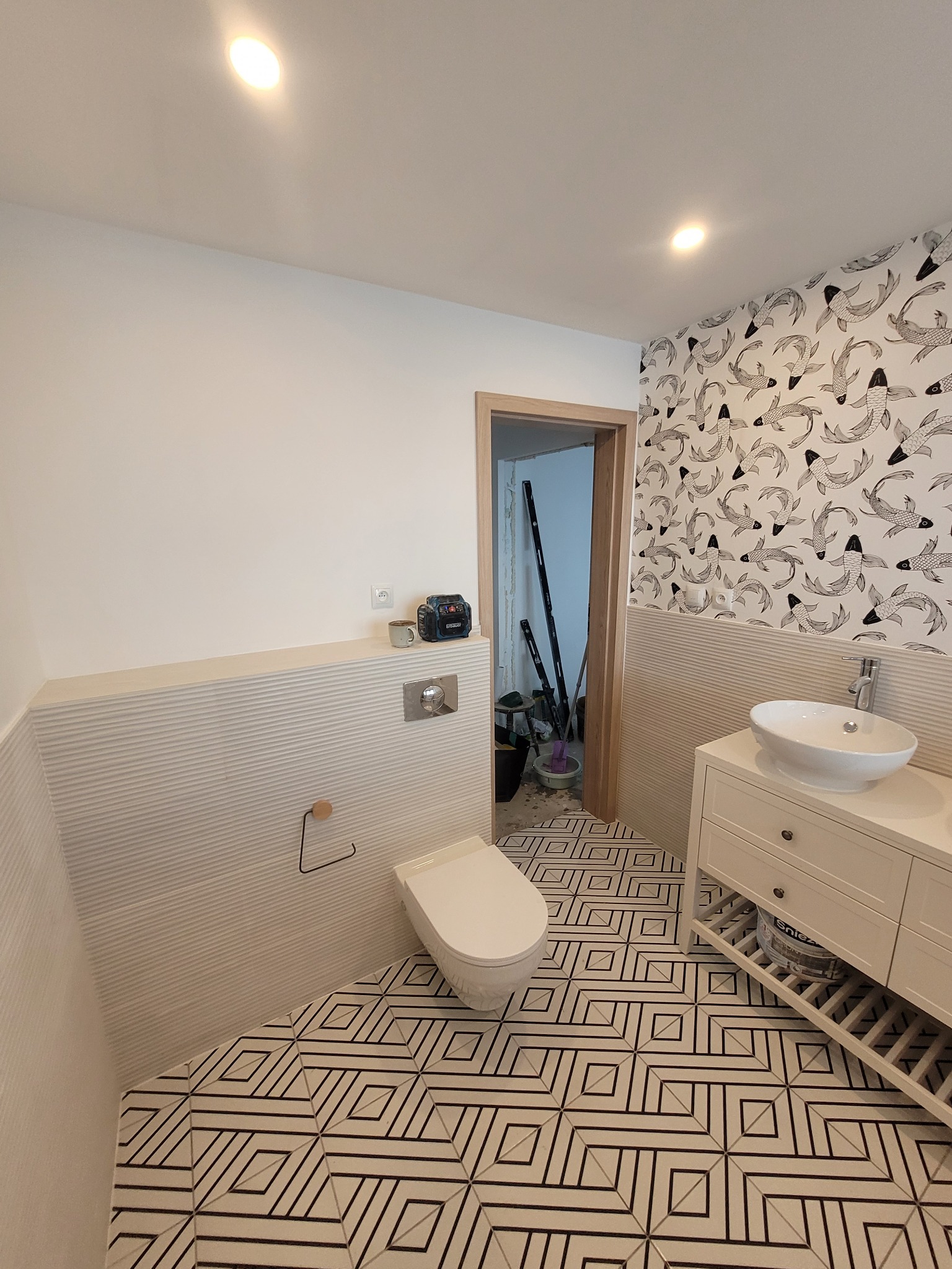 Nowoczesna łazienka z geometryczną podłogą, białą armaturą i tapetą w ryby, widok na otwarte drzwi z narzędziami w tle.