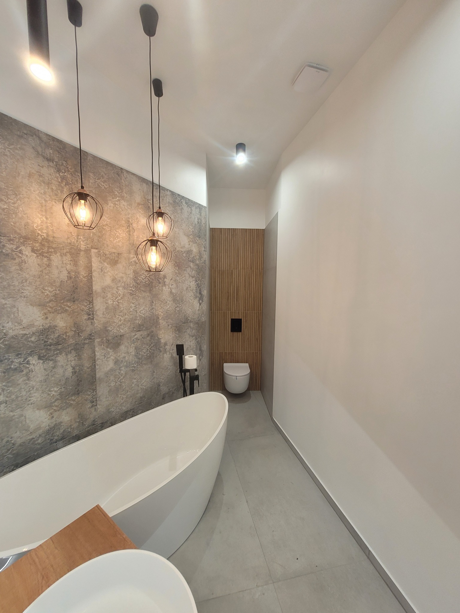 Nowoczesna łazienka z wolnostojącą wanną, betonową ścianą i wiszącymi lampami w stylu industrialnym, akcentowana drewnianą ścianką i minimalistyczną toaletą.