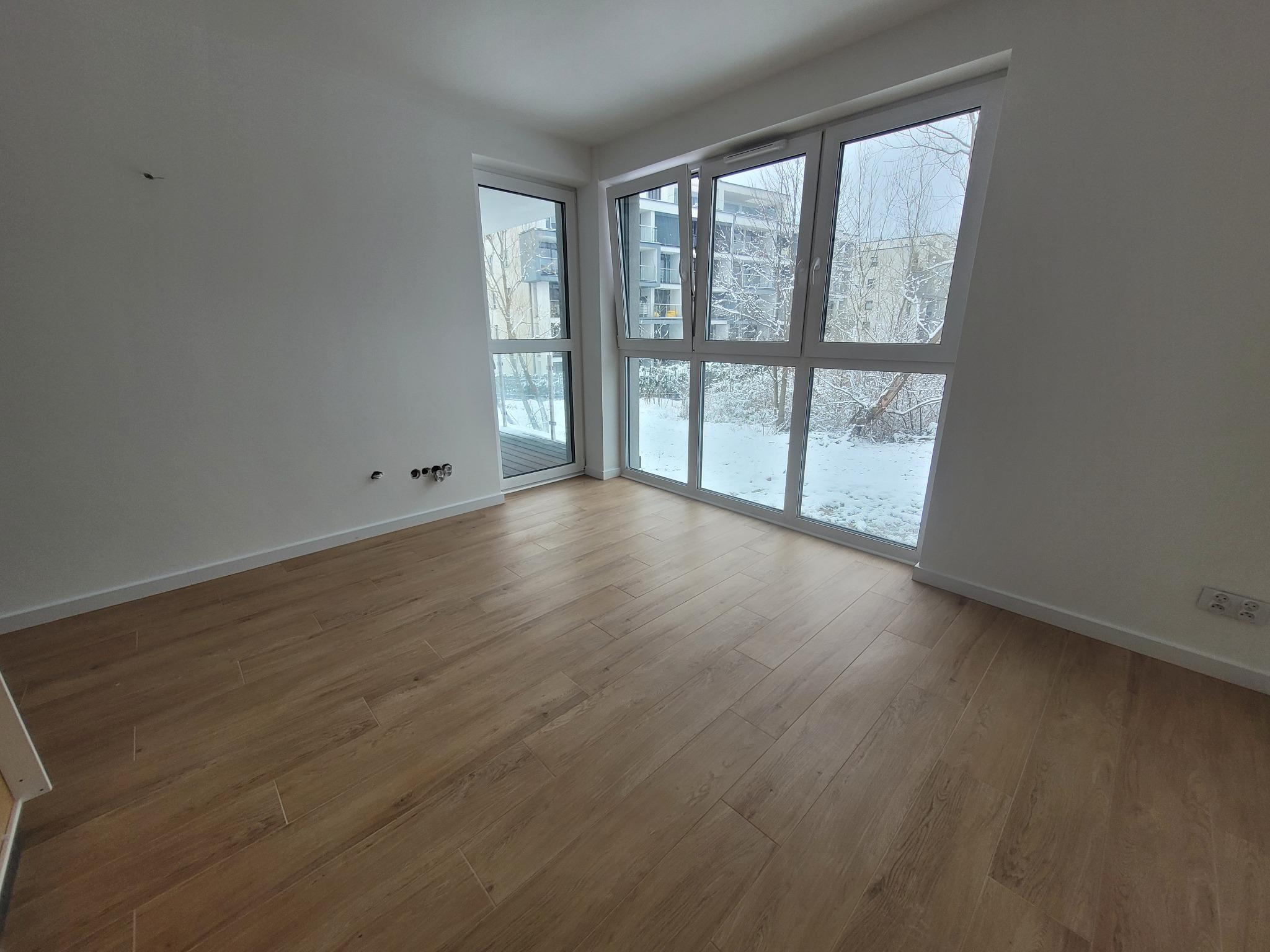 Wnętrze pustego, jasnego pokoju z drewnianą podłogą i dużymi oknami z widokiem na zimowy krajobraz.