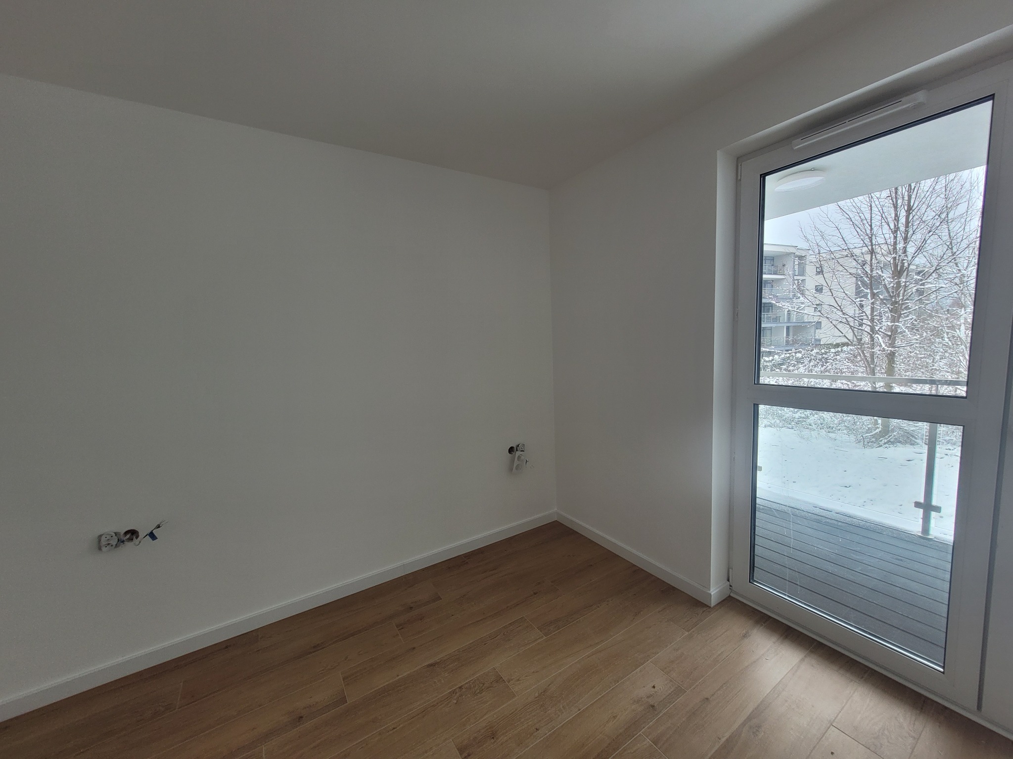 Pusty pokój z białymi ścianami, drewnianą podłogą i wyjściem na balkon zasypany śniegiem, widoczne wystające kable elektryczne.