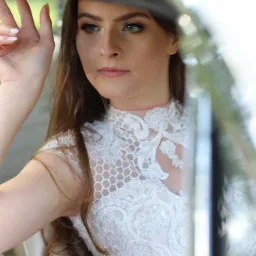 Portret kobiety w białej, koronkowej sukni, siedzącej w samochodzie z jasną tapicerką, z ręką opartą na framudze okna.