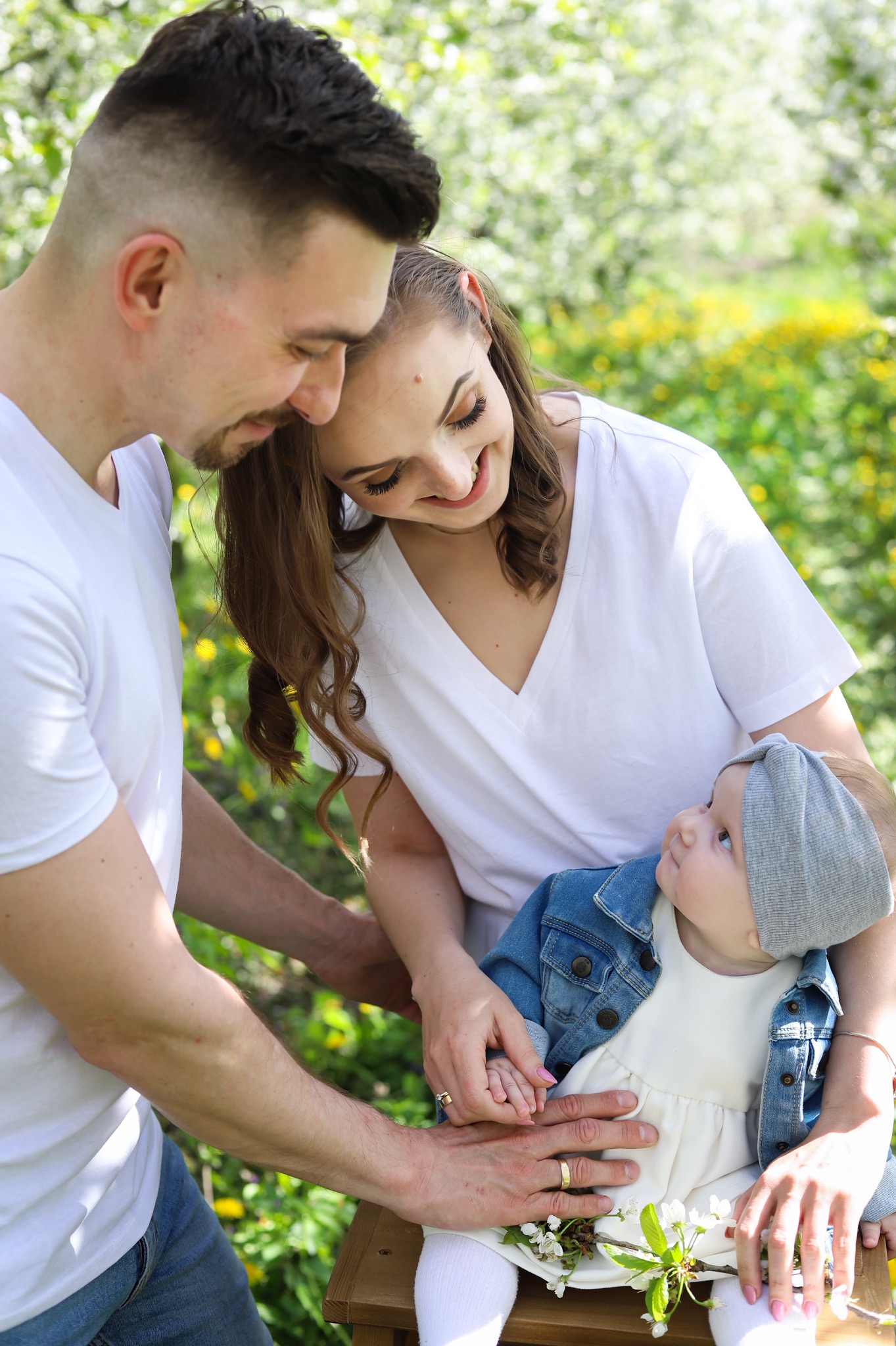 Rodzina: tata z modną fryzurą, mama z delikatnym makijażem i uśmiechnięta córeczka w dżinsowej kurtce, siedząca na kolanach, na tle kwitnących drzew.