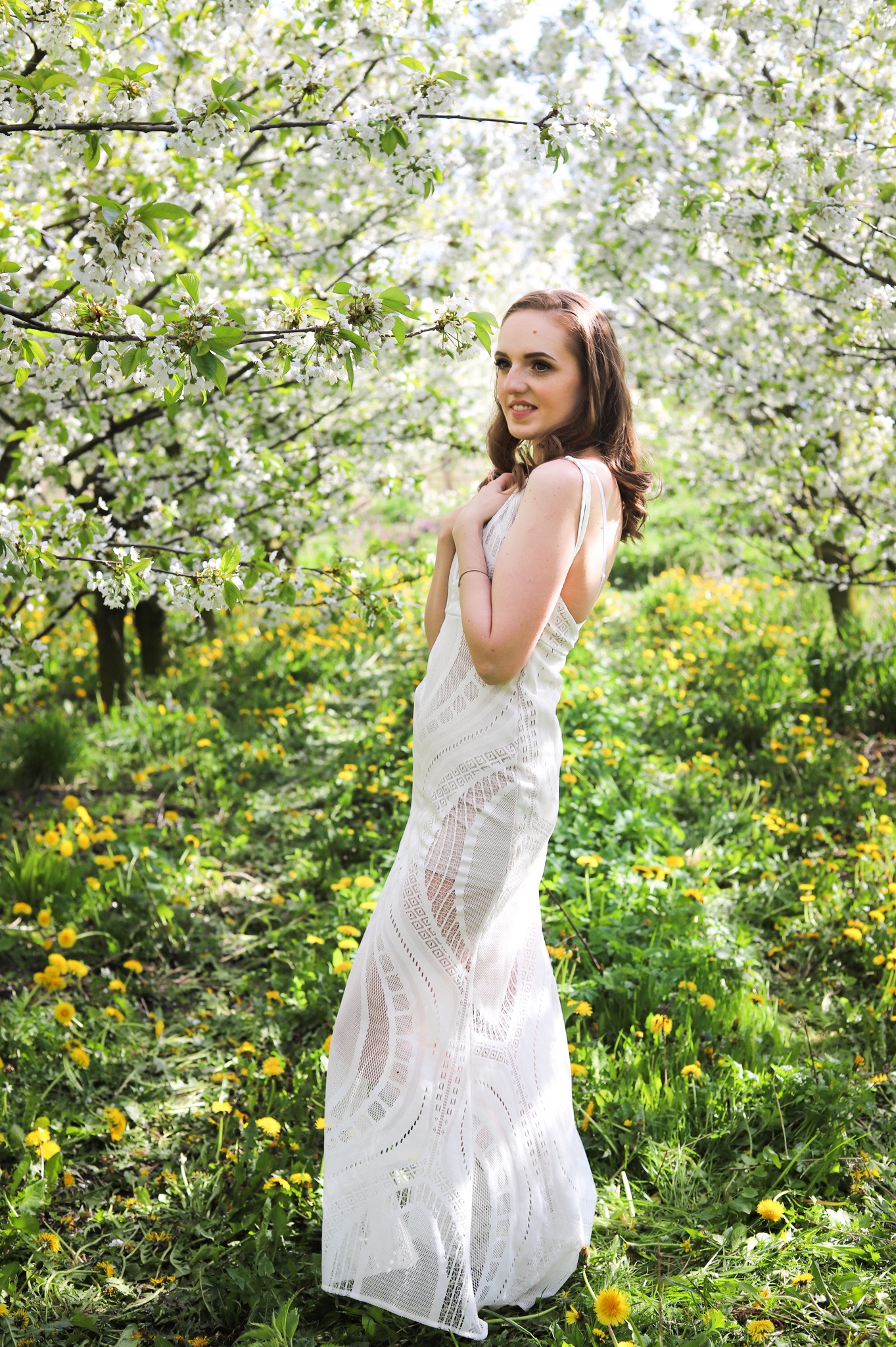 Kobieta w zwiewnej białej sukience pozuje w sadzie kwitnących drzew, otoczona zielenią i żółtymi kwiatami.