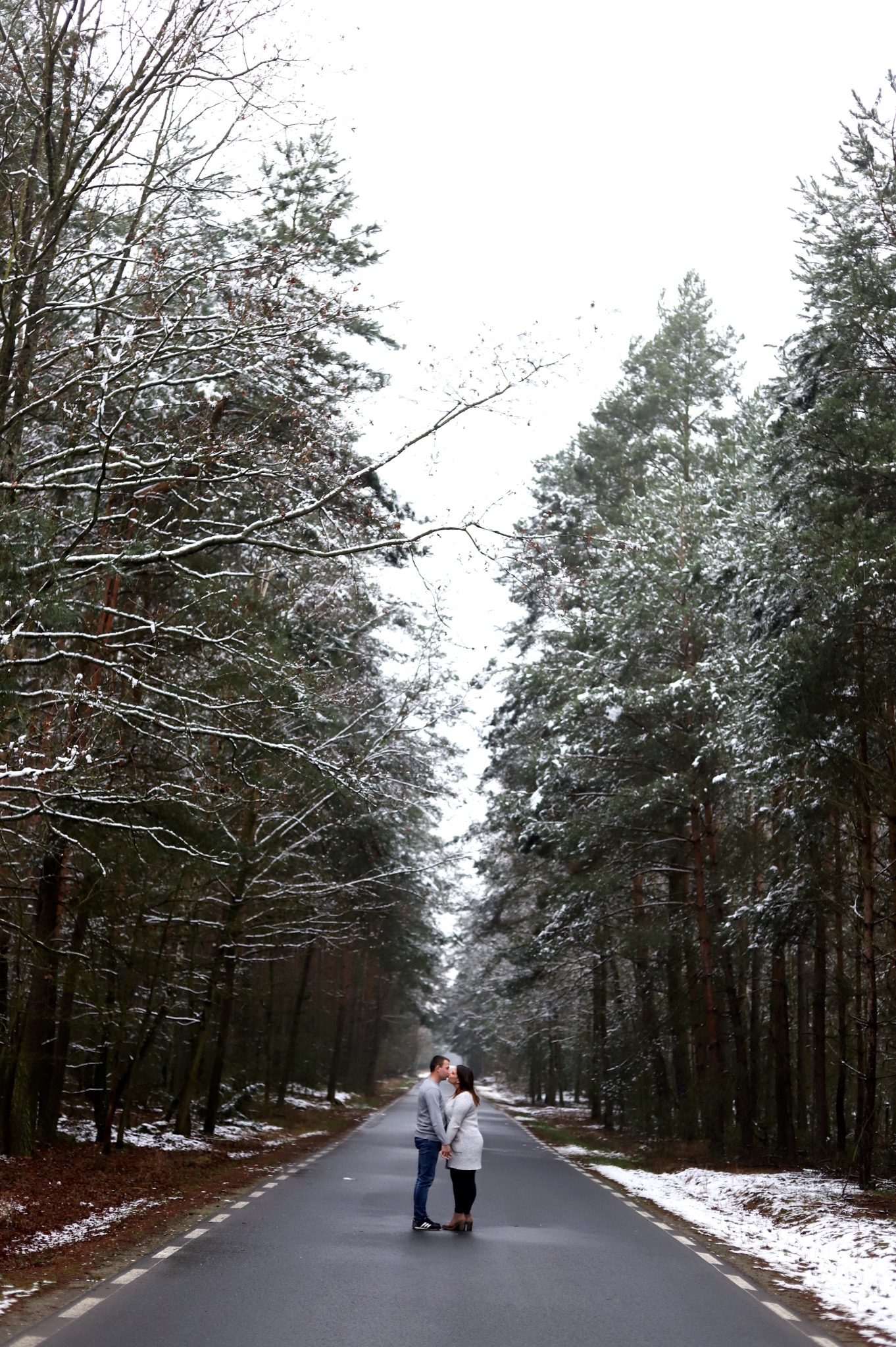 Para trzymająca się za ręce, stojąca na asfalcie w lesie zimą, drzewa pokryte śniegiem.
