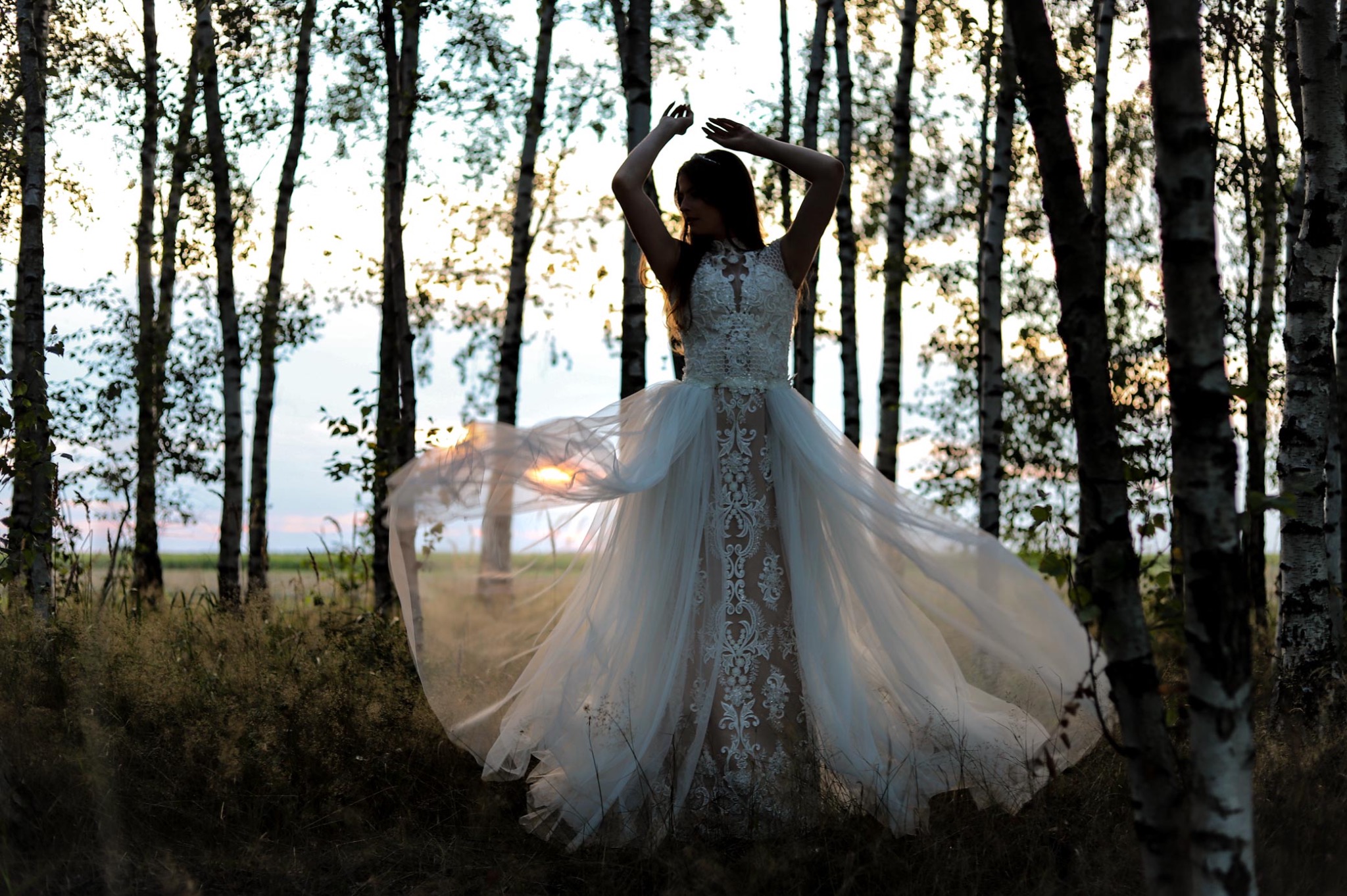 Kobieta w białej, koronkowej sukni z tiulowym dołem pozuje z podniesionymi rękami w lesie brzozowym o zmierzchu.