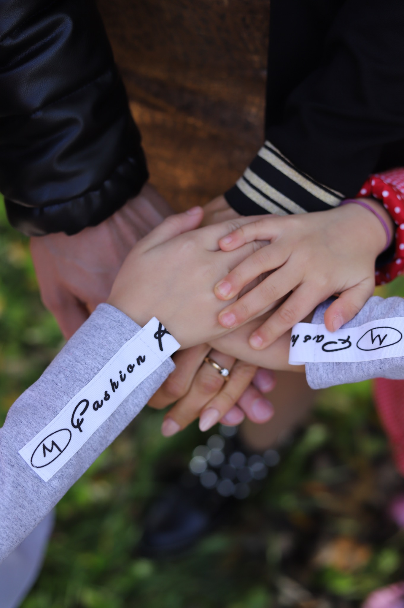 Dłonie członków rodziny ułożone jedna na drugiej, widoczny pierścionek zaręczynowy na palcu kobiety, ubrania z napisami Fashion, na tle zieleni.