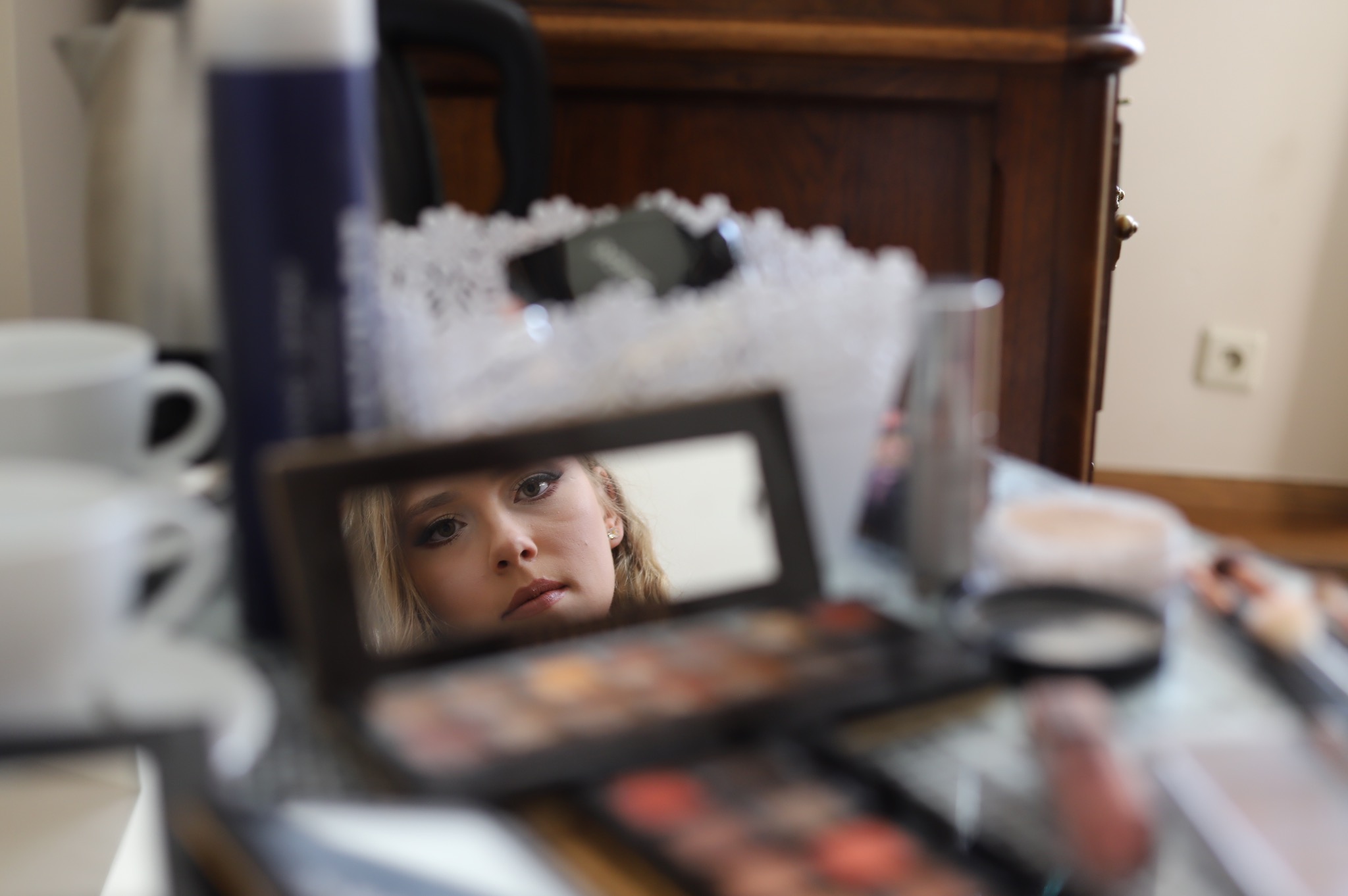 Odbicie w lusterku twarzy kobiety w trakcie makijażu, paleta cieni i kosmetyki na stole, rozmyte tło z meblami.