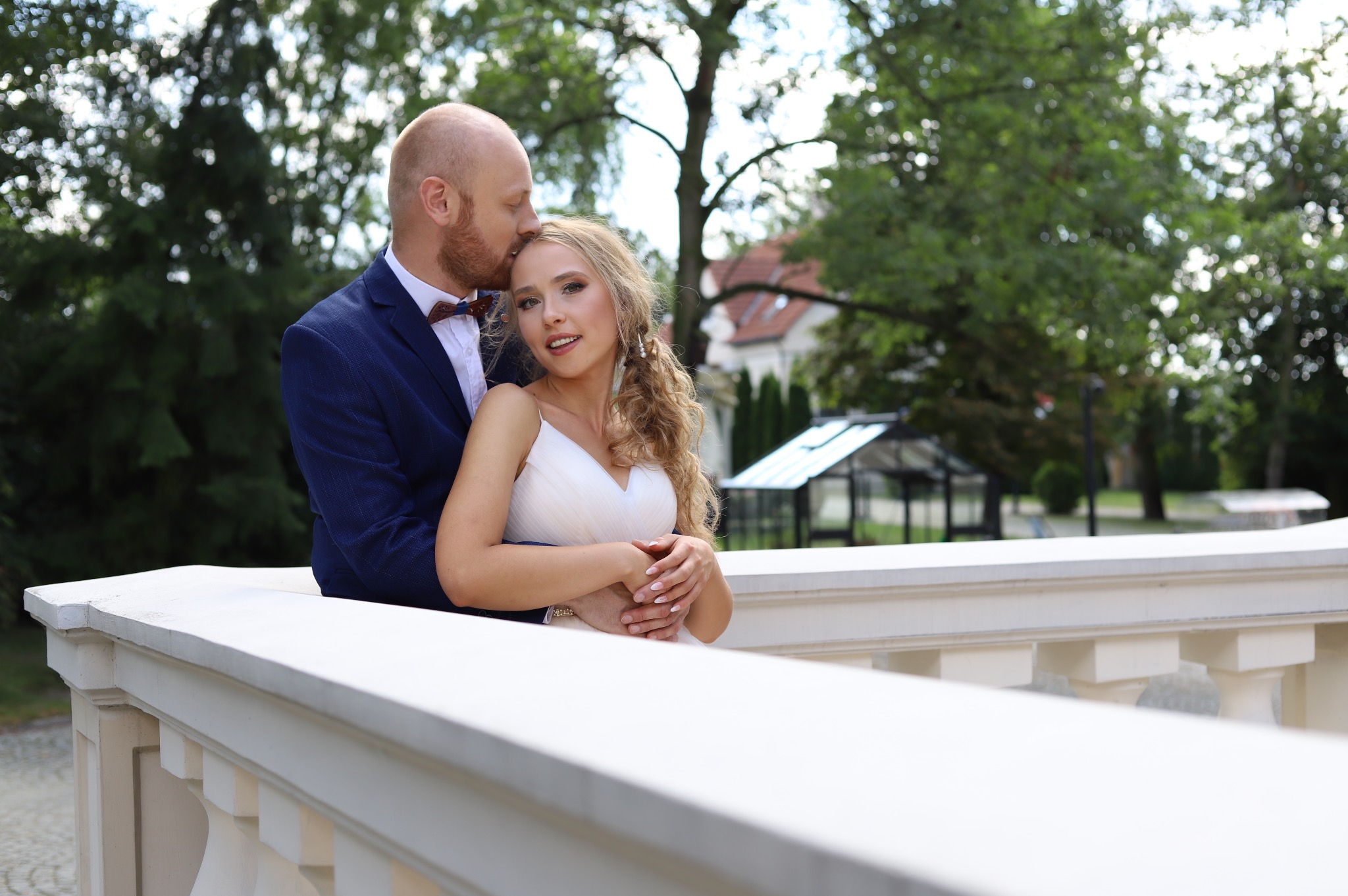 Para narzeczeńska: mężczyzna w granatowej marynarce całuje kobietę w białej sukience na tle zieleni i białej balustrady.