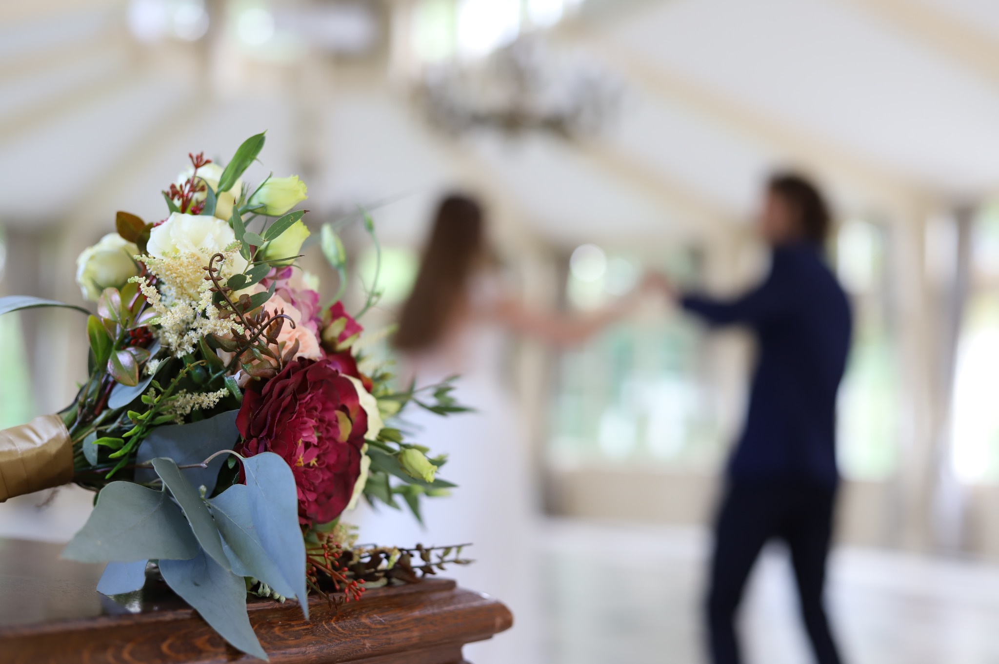 Bukiet ślubny z burgundowymi i białymi kwiatami na drewnianym stole, para tańcząca w tle w jasnym wnętrzu.