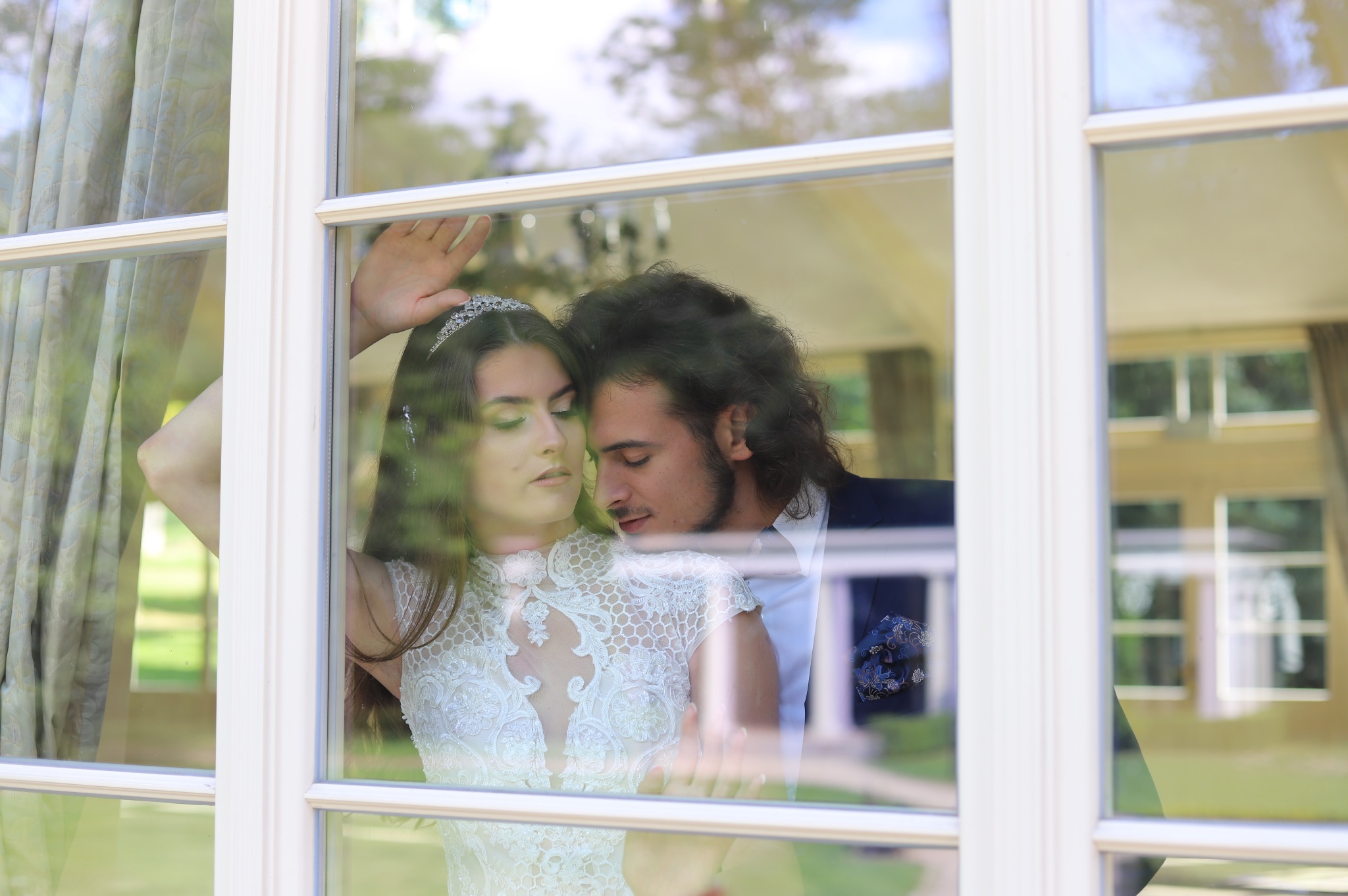 Para narzeczonych w objęciach, widziana przez okno z białymi ramami, ona w koronkowej sukni i diademie, on w ciemnym garniturze z wzorzystą poszetką, delikatne światło.