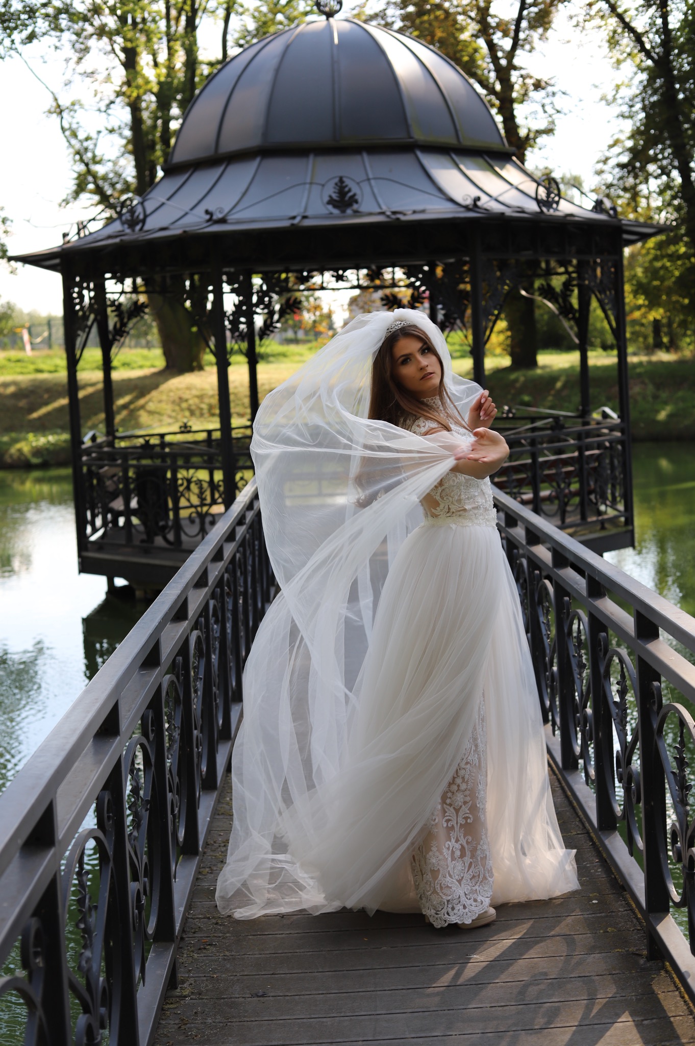 Kobieta w białej sukni pozuje na drewnianym moście z metalową balustradą, z wiatrem unoszącym jej welon, w tle altana nad wodą i drzewa.