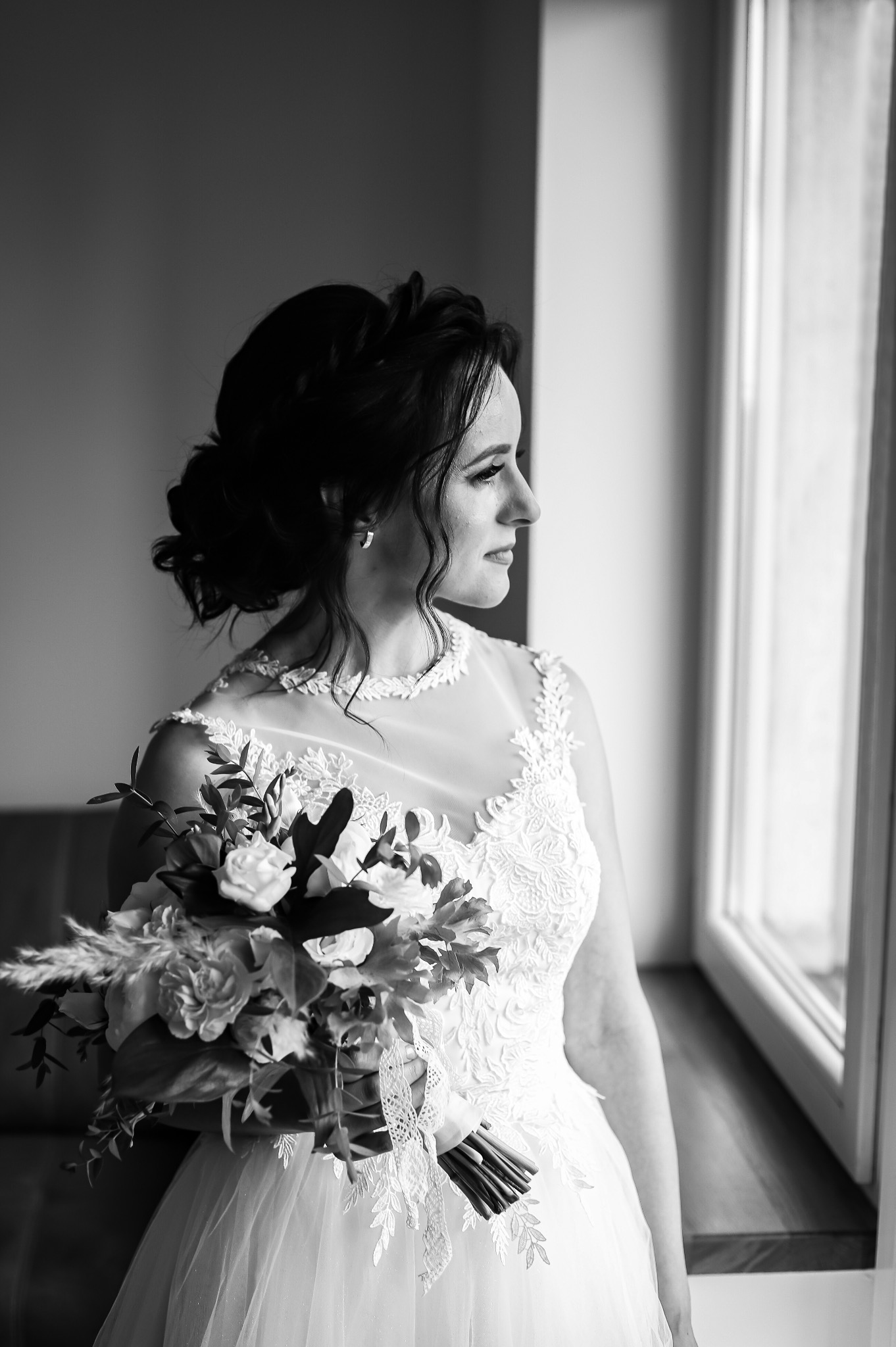 Panna młoda w koronkowej sukni spogląda przez okno, trzymając bukiet z jasnych kwiatów i dekoracyjną wstążką, zdjęcie czarno-białe.