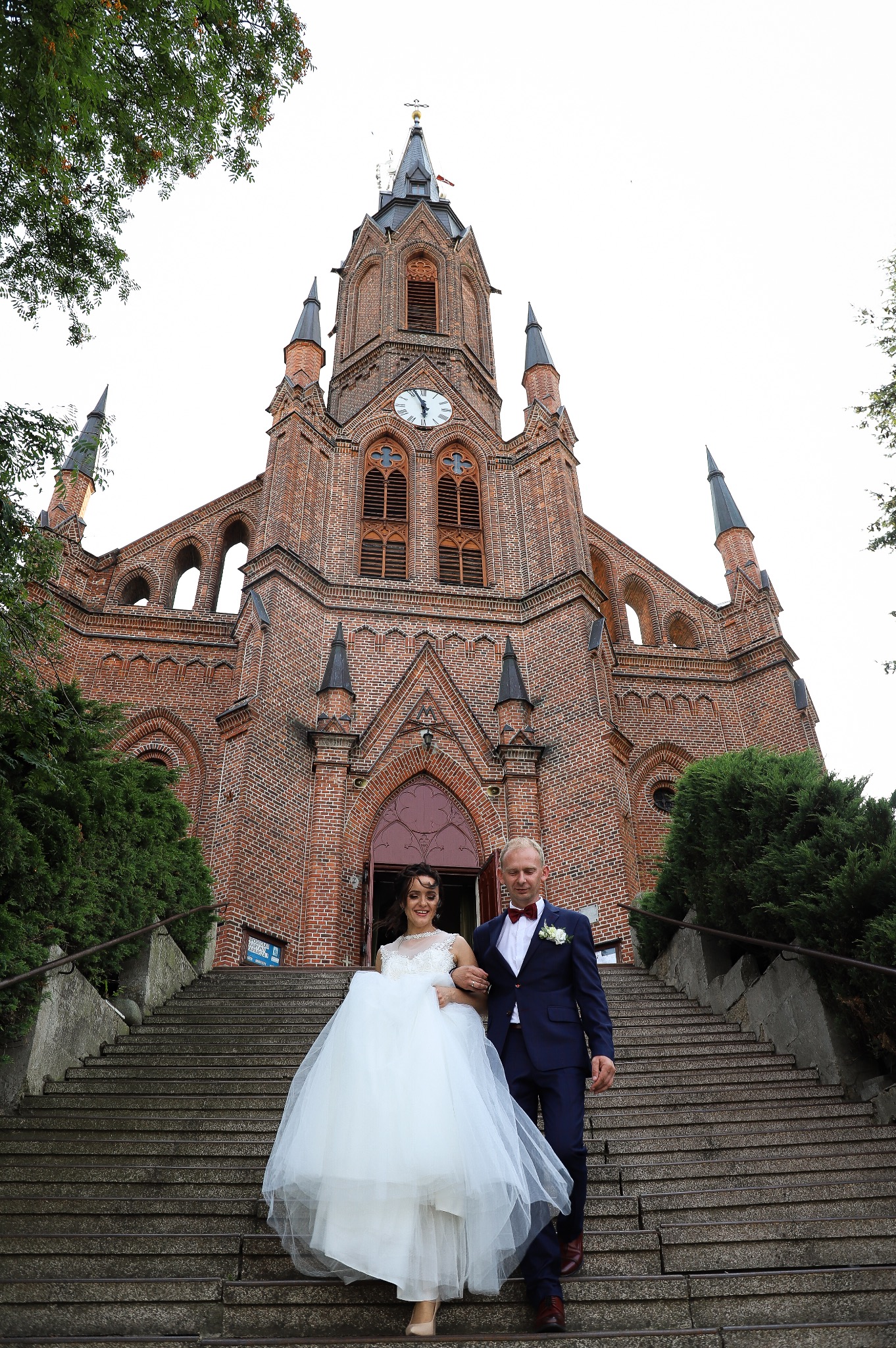 Para młoda schodzi po schodach kościoła z czerwonej cegły w dniu ślubu; panna młoda w białej sukni, pan młody w granatowym garniturze z czerwoną muszką.