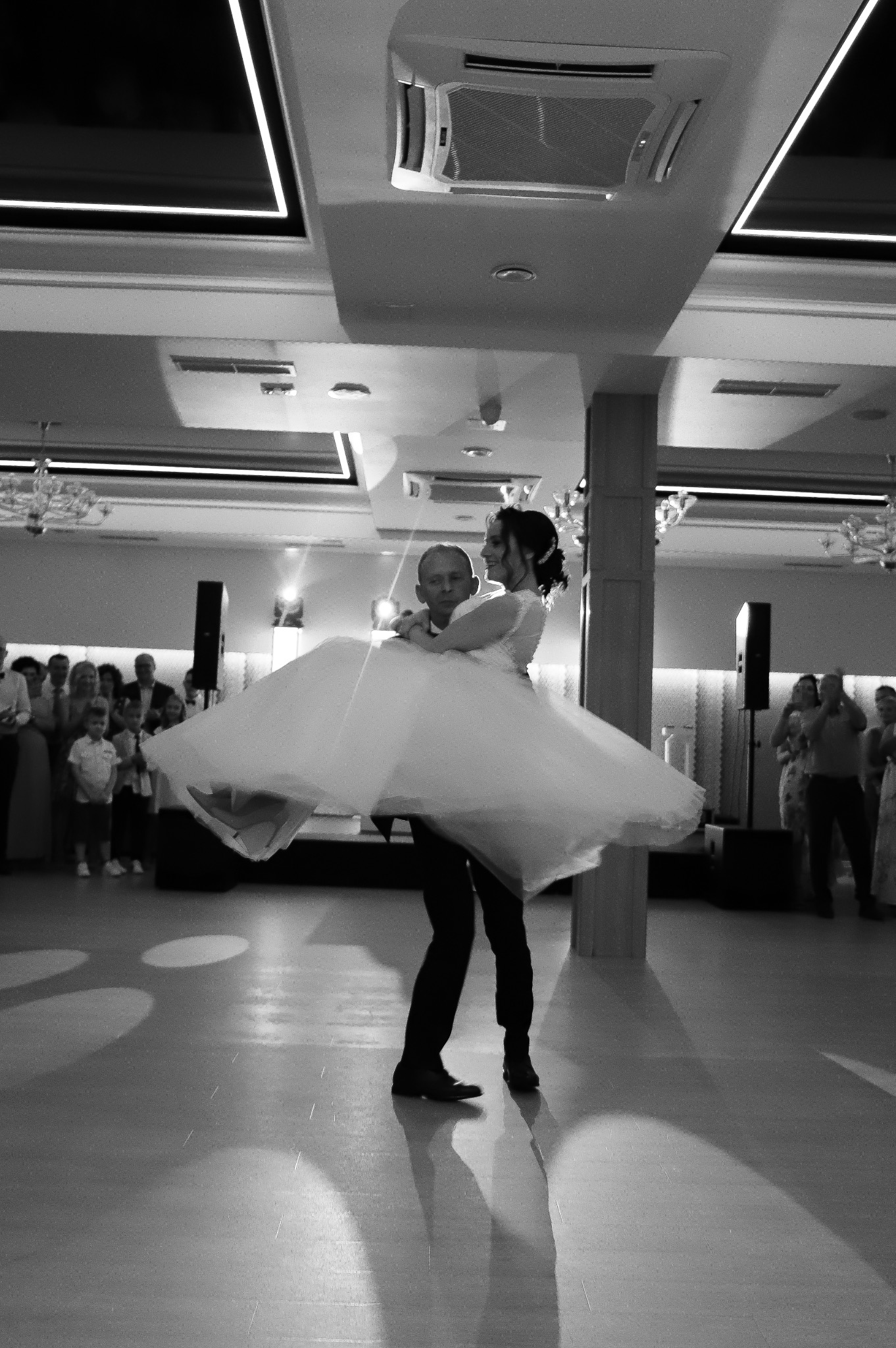 Czarno-białe zdjęcie: Pan młody trzyma pannę młodą w ramionach podczas pierwszego tańca na sali balowej, goście obserwują w tle.