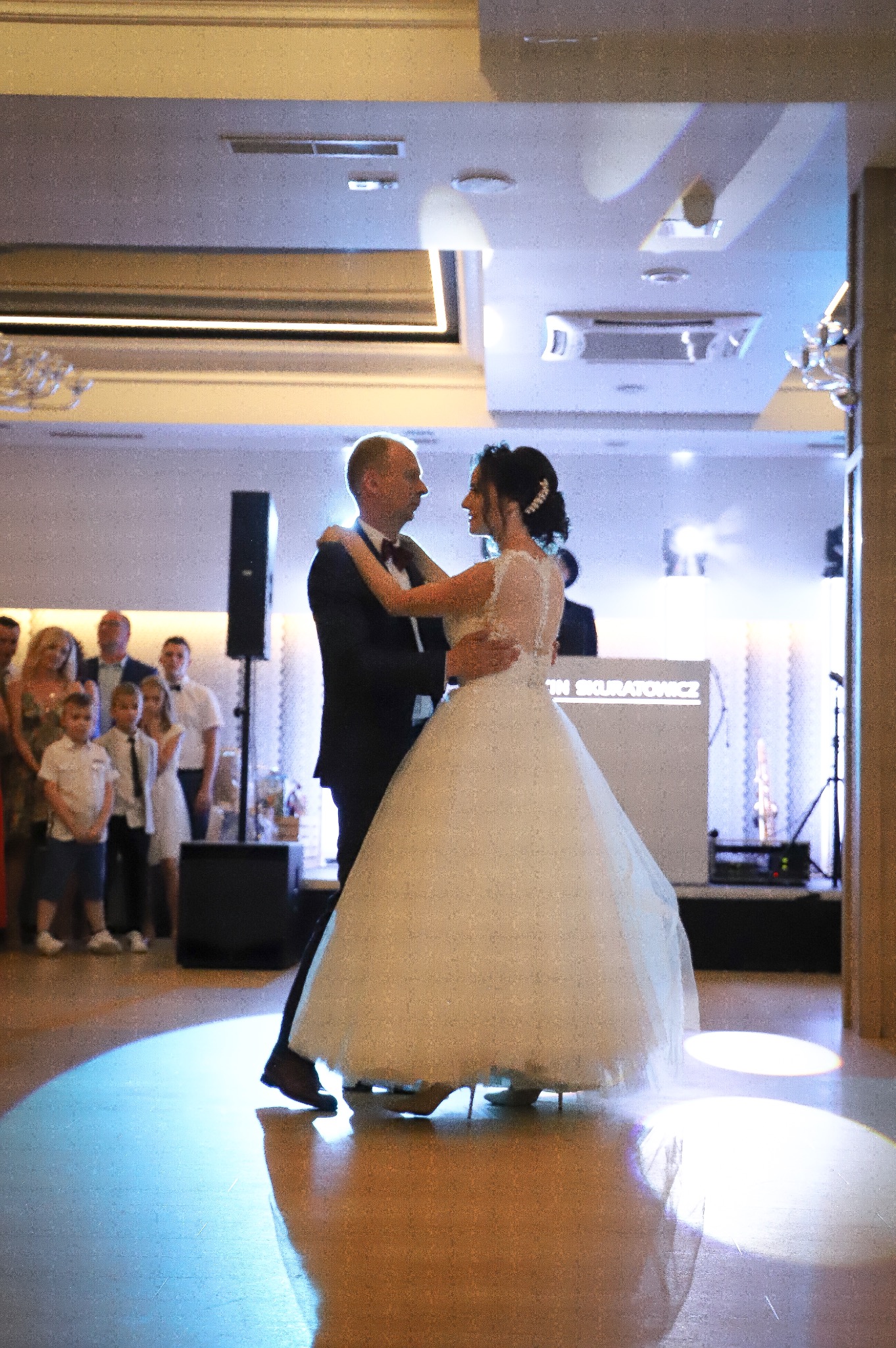 Para młoda tańczy pierwszy taniec w sali weselnej, goście obserwują w tle, reflektory oświetlają parkiet.