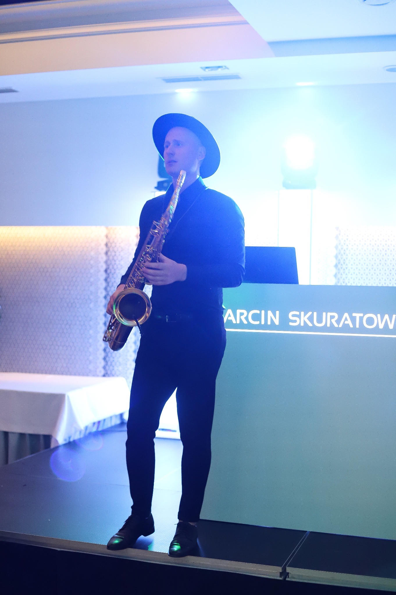 Saksofonista w czarnym stroju i kapeluszu na scenie z podświetlonym napisem 'ARCI SKURATOW'