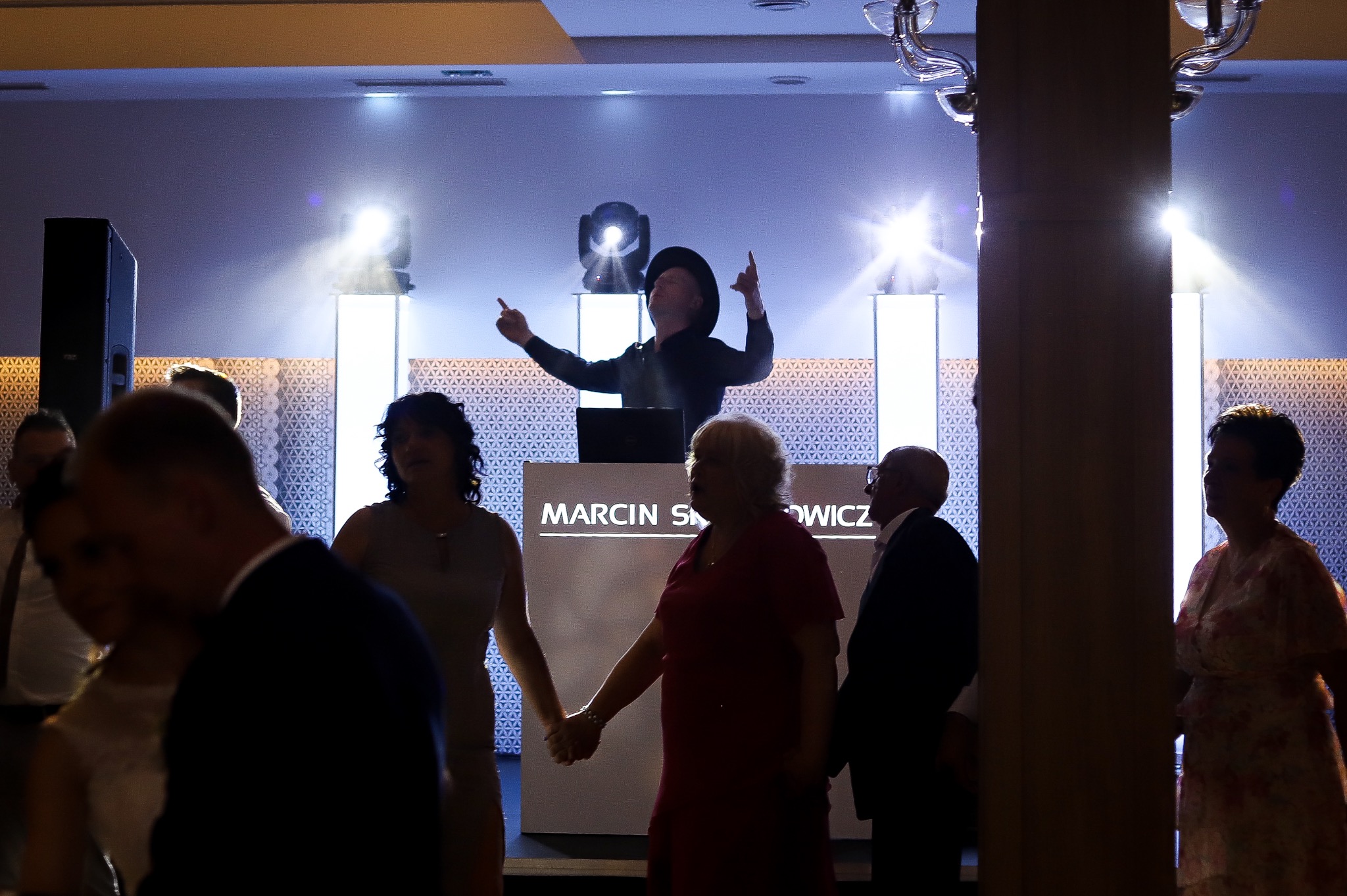 DJ w czarnym kapeluszu z podniesionymi rękami za konsoletą z napisem Marcin S. podczas imprezy, oświetlony reflektorami, z widocznymi gośćmi w tle.