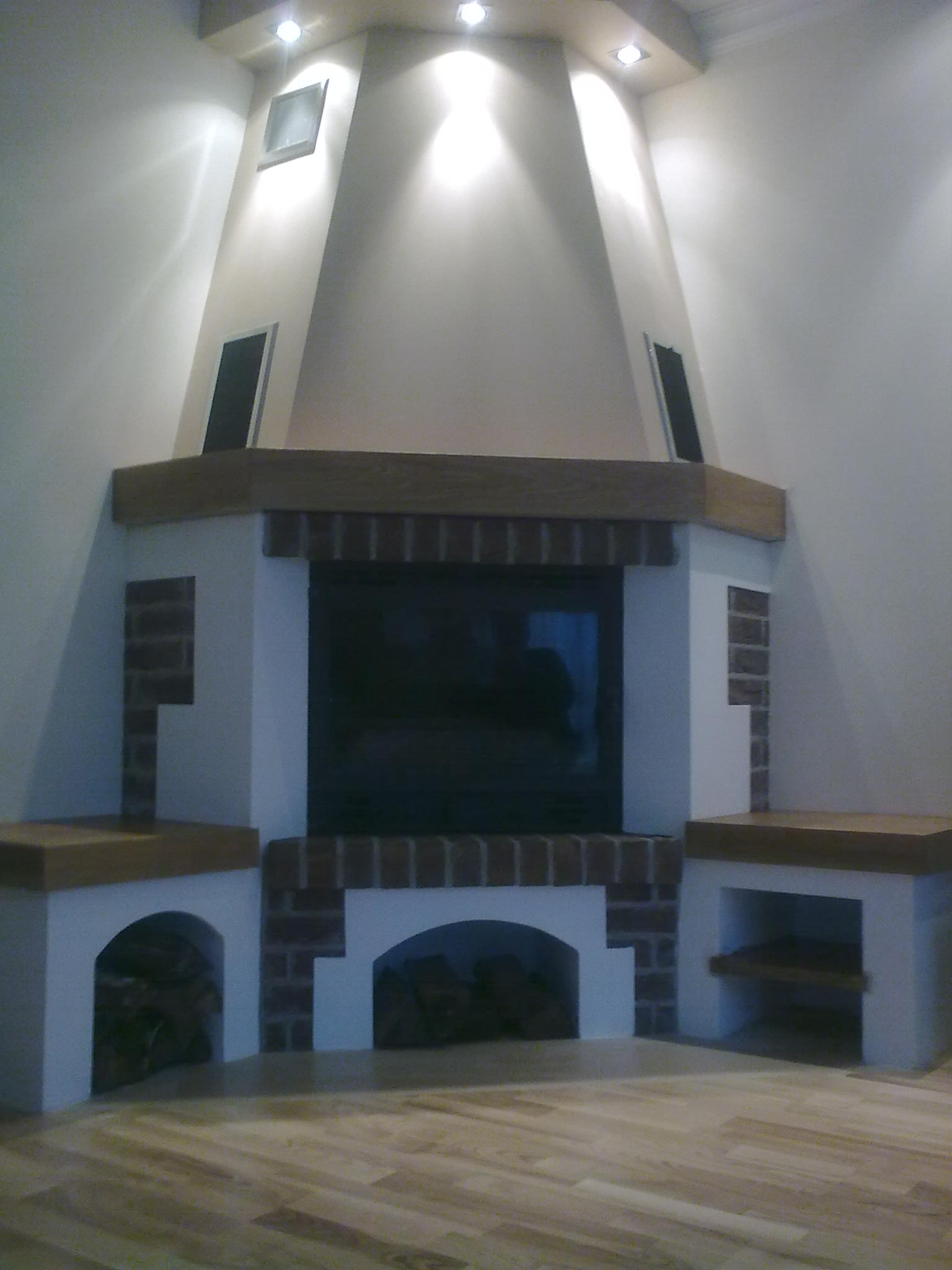 Komin z ceglanymi elementami i drewnianymi półkami na drewno, oświetlony halogenami, na tle parkietu.