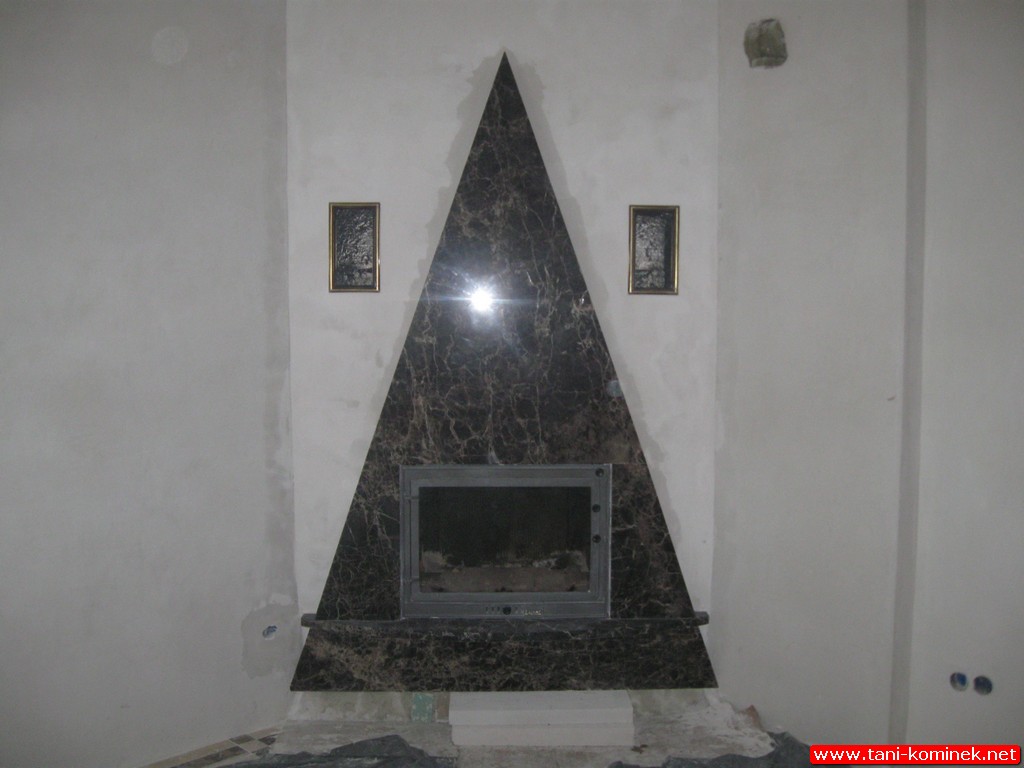 Kominek z obudową z ciemnego marmuru w kształcie trójkąta, z dwoma małymi obrazkami po bokach, w trakcie montażu, widoczne niedociągnięcia w wykończeniu ściany.