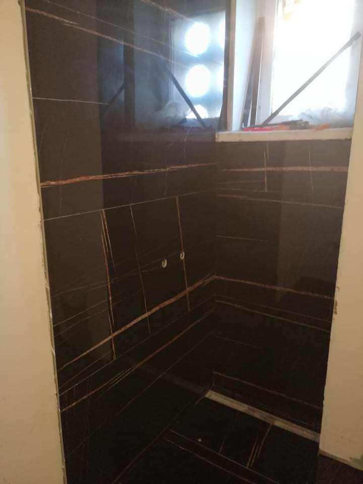 Wykończona kabina prysznicowa z ciemnymi płytkami imitującymi marmur ze złotymi żyłami, widoczne otwory na baterię, okno w tle z częściowo zakrytym widokiem.