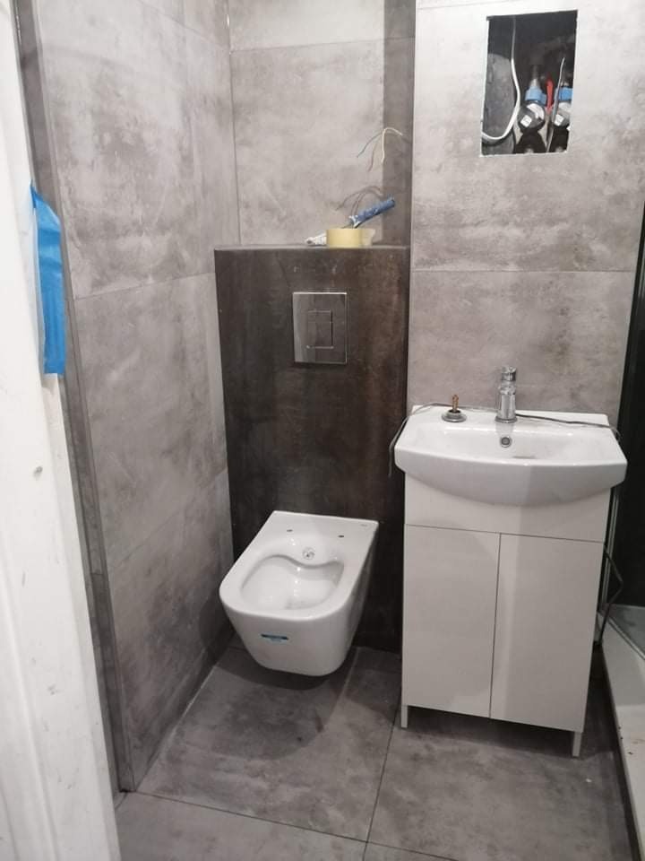 Niedokończona łazienka z szarymi płytkami imitującymi beton, białą umywalką z szafką, bidetem i otwartą instalacją hydrauliczną w ścianie.