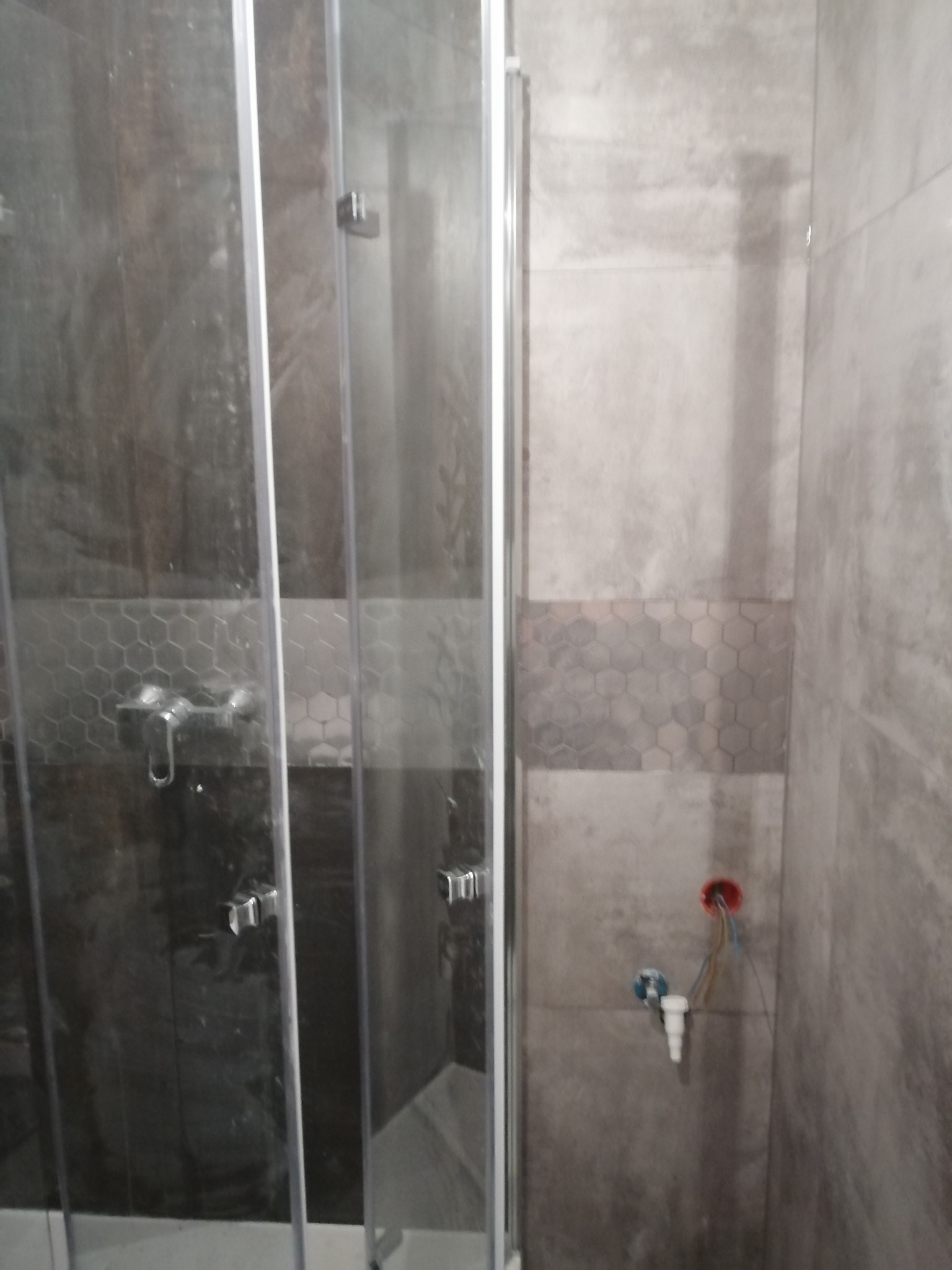 Szara łazienka z kabiną prysznicową, widoczne płytki imitujące beton i dekoracyjny pas z heksagonów, niezamaskowane przyłącze wodno-kanalizacyjne oraz instalacja elektryczna.