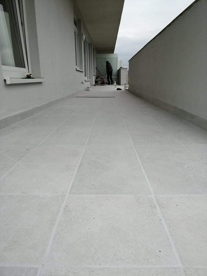 Długi balkon wyłożony jasnoszarą, kwadratową terakotą; w tle widoczna osoba sprzątająca po zakończonych pracach.