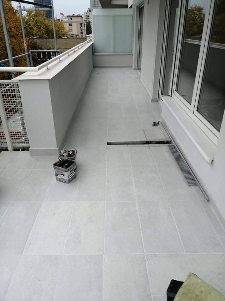 Wyłożony jasnymi płytkami balkon z odpływem liniowym, widoczne narzędzia w wiaderku, balustrada z metalowymi elementami, widok na budynki w tle.