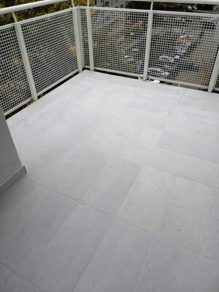 Jasnoszary, kwadratowy gres na podłodze balkonu z białą, metalową balustradą o siatkowej strukturze. Widok na drzewa i zabudowę miejską w tle.