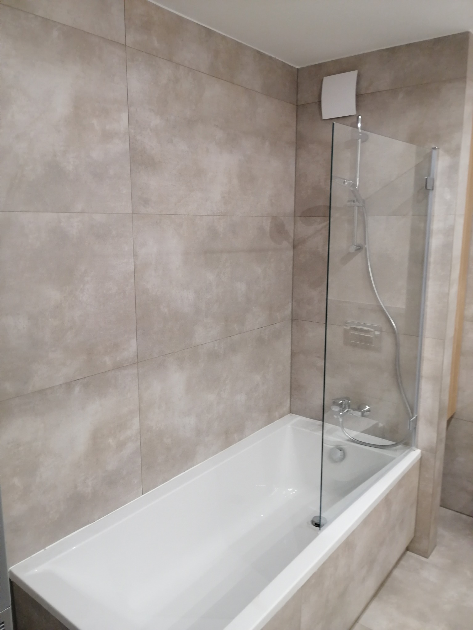 Wykończona łazienka w Warszawie z wanną i szklanym parawanem nawannowym, ściany wyłożone beżowymi, kwadratowymi płytkami imitującymi kamień.