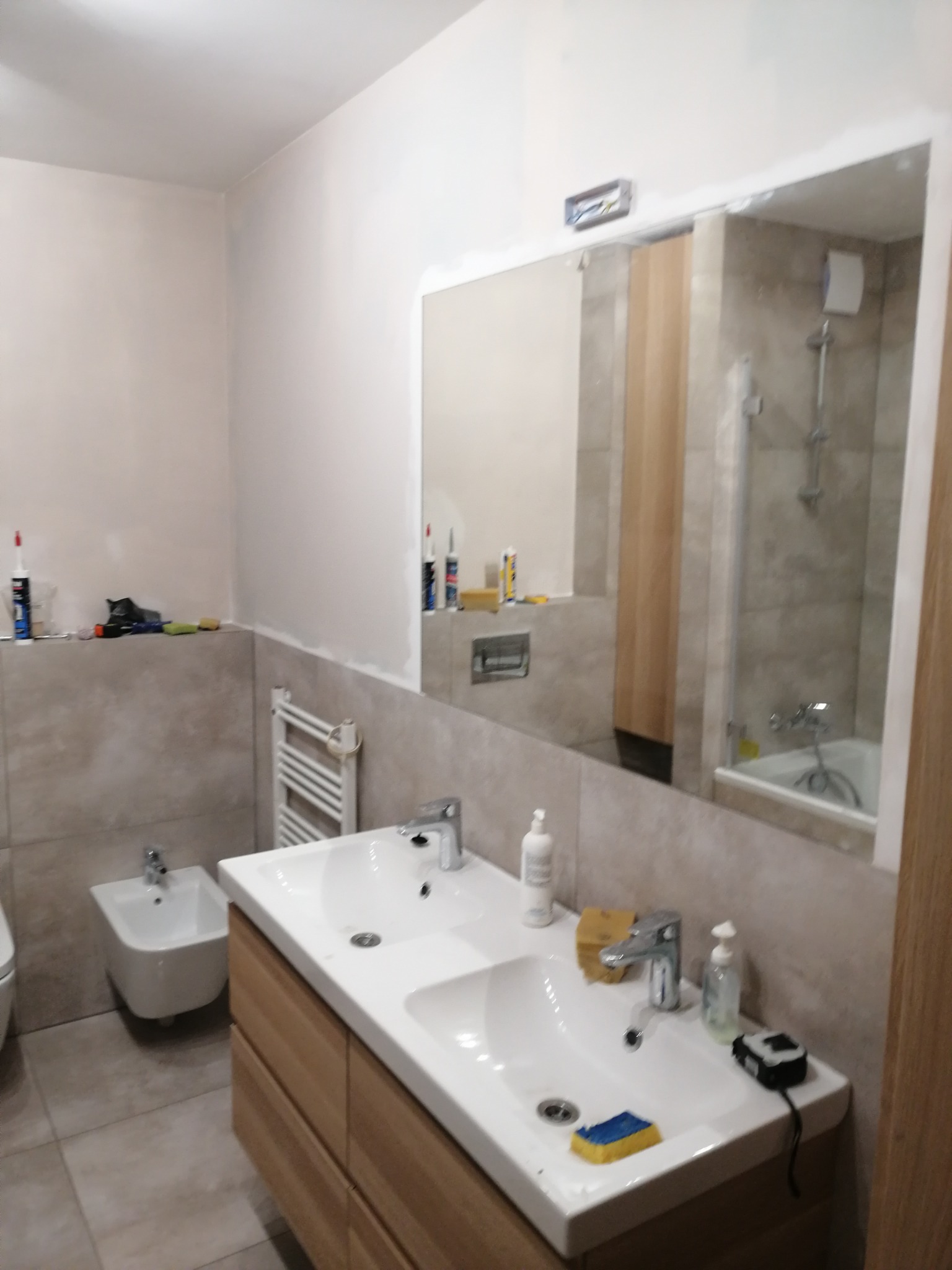 Łazienka w trakcie remontu: widoczne dwie umywalki z szafkami z jasnego drewna, bidet, grzejnik drabinkowy, lustro z odbiciem kabiny prysznicowej, narzędzia i materiały budowlane na półce z płytek.