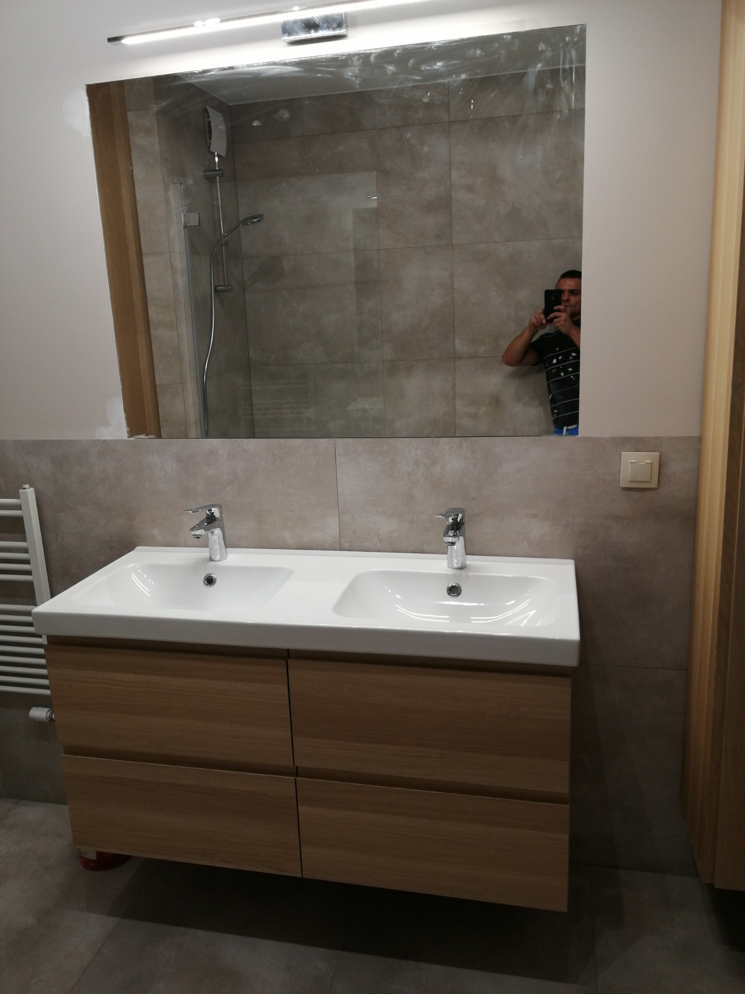 Nowoczesna łazienka z podwójną umywalką na drewnianej szafce, szare płytki na ścianach i w kabinie prysznicowej, odbicie fotografa w lustrze.