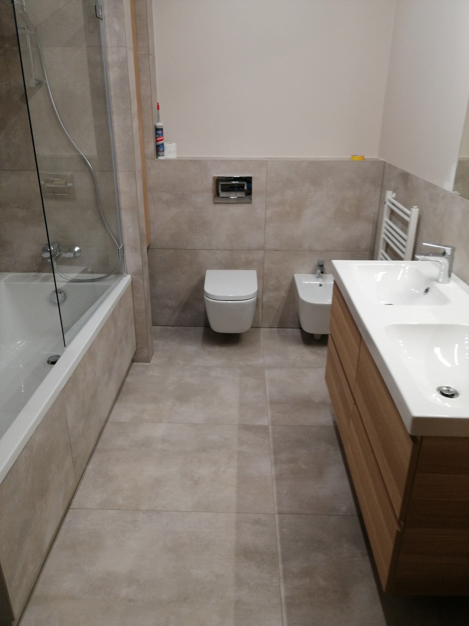 Wykończona łazienka z beżowymi płytkami, wiszącą toaletą i bidetem, oraz umywalką z drewnianą szafką. Na ścianie widoczny grzejnik drabinkowy.