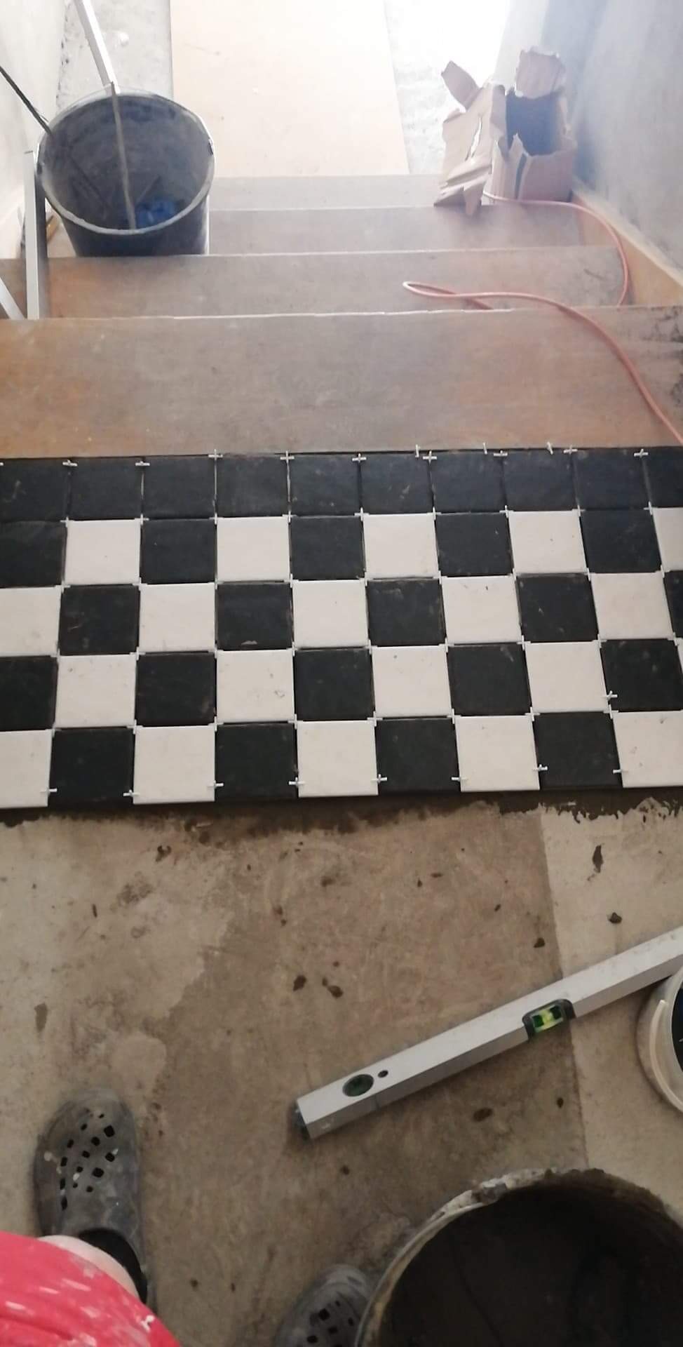 Układanie czarno-białej, szachownicowej mozaiki z płytek na podłodze przy schodach, widoczne krzyżyki dystansowe, poziomica i wiadro z zaprawą.