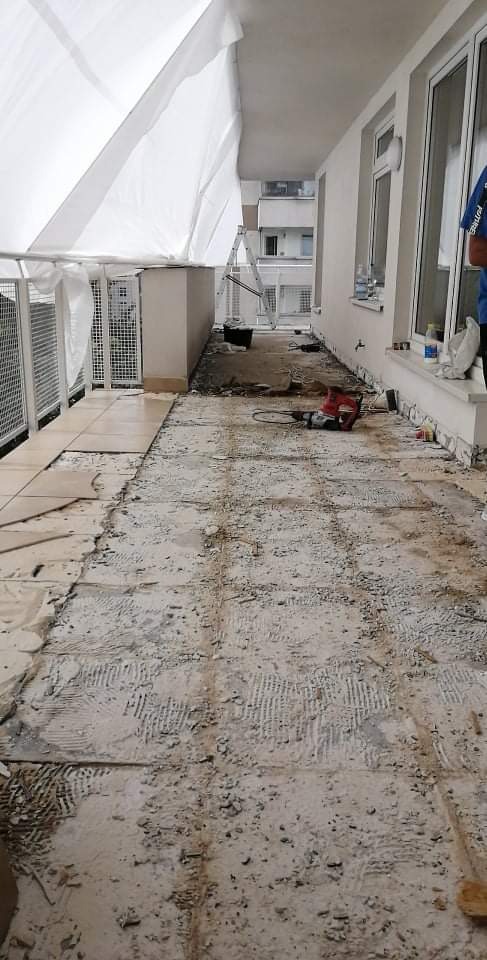 Demontaż starej terakoty na balkonie z użyciem młota udarowego, widoczne częściowo położone nowe płytki, zabezpieczenie folią malarską nad głową, drabina oparta o balustradę.