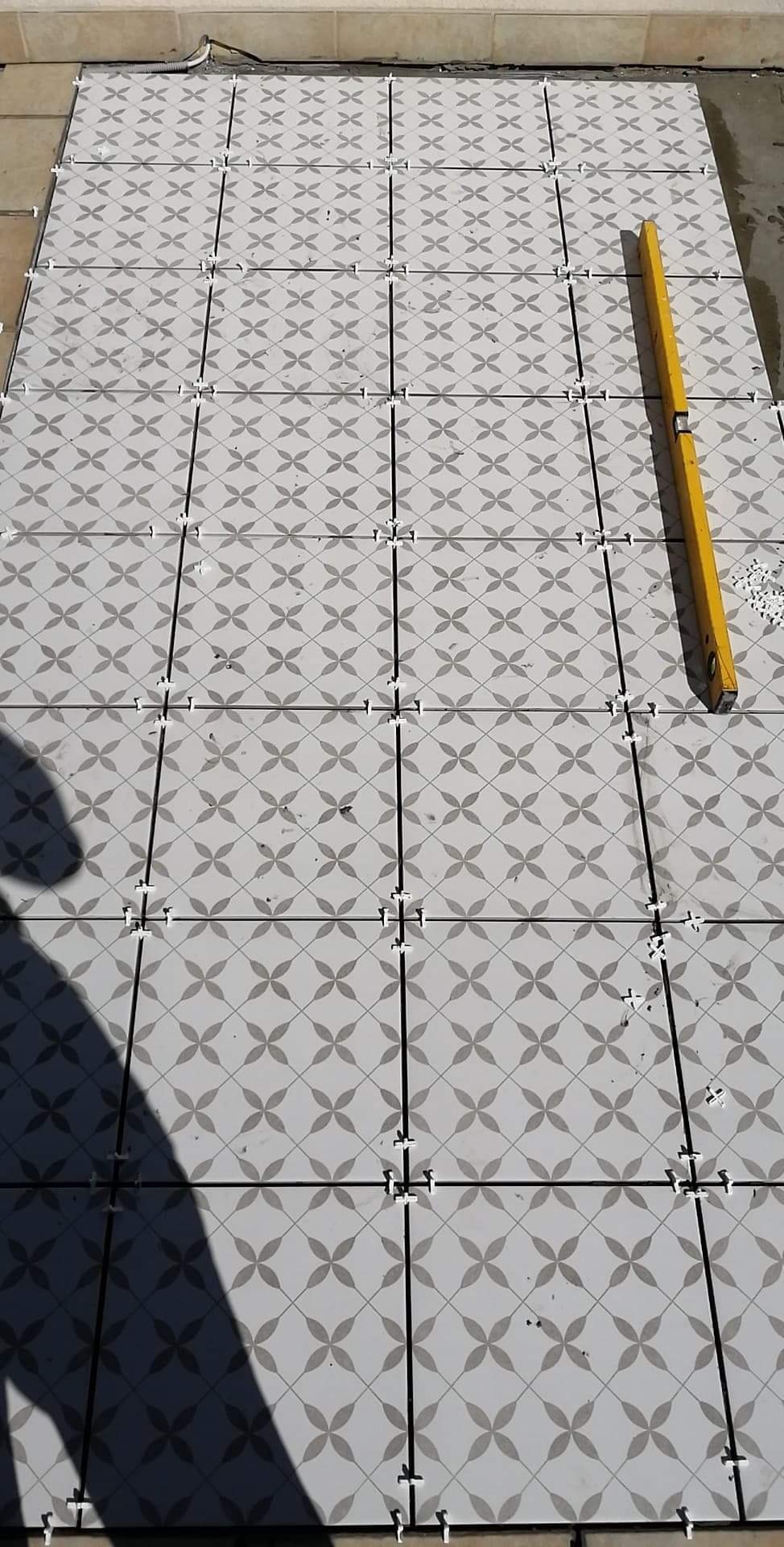 Układanie jasnych, kwadratowych płytek z szarym wzorem w kształcie czterolistnej koniczyny, widoczne krzyżyki dystansowe i poziomica w trakcie prac.