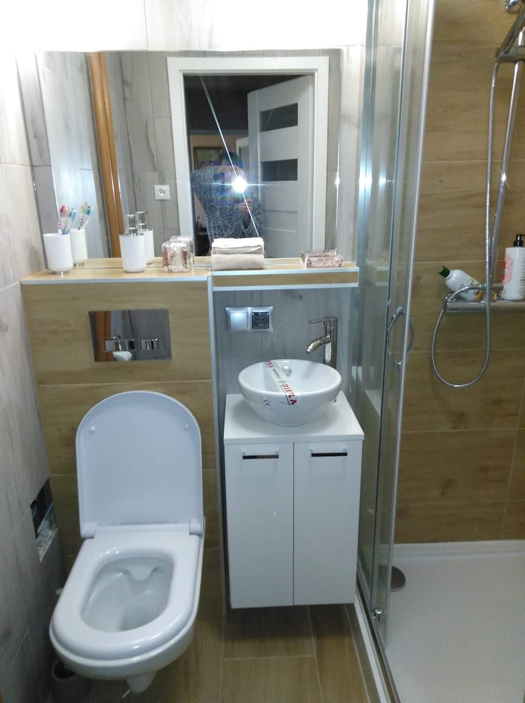 Nowoczesna łazienka z beżowymi płytkami imitującymi drewno, wisząca toaleta z podtynkową spłuczką, umywalka nablatowa na białej szafce oraz kabina prysznicowa z widocznym zestawem natryskowym.