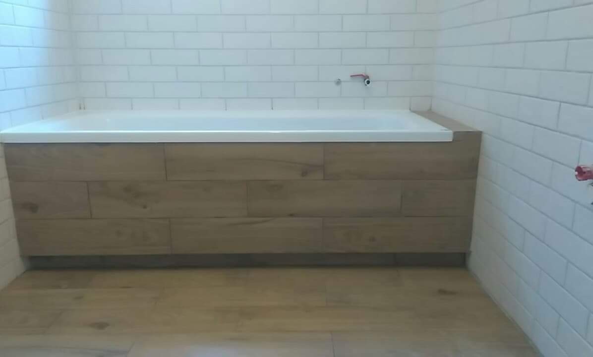 Wykończona łazienka w Warszawie z wanną obudowaną panelami imitującymi drewno, białe, prostokątne płytki na ścianach i podłodze.