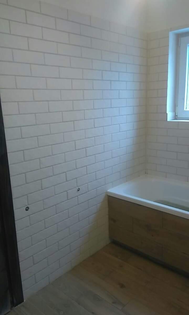 Białe, prostokątne płytki ułożone na ścianie w łazience, z widocznymi dwoma otworami na rurę oraz fragmentem drewnopodobnej obudowy wanny i drewnianej podłogi.