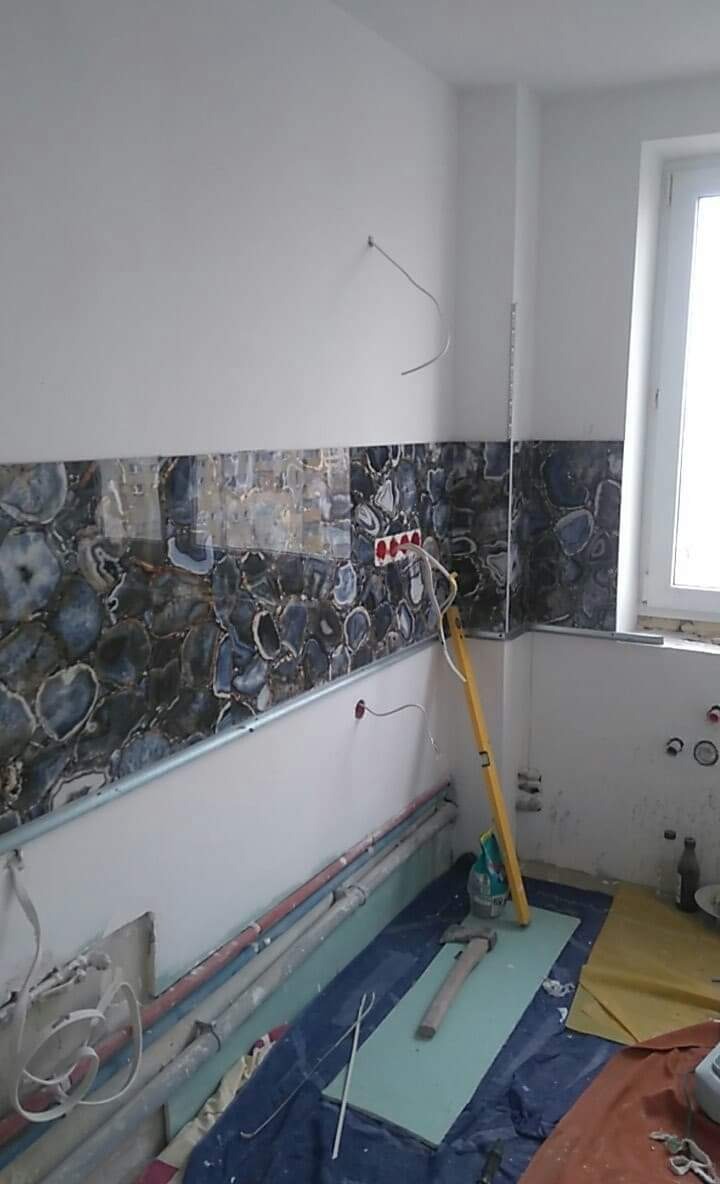 Fragment ściany w trakcie prac wykończeniowych, z położonym pasem dekoracyjnych płytek imitujących agat, widoczne wystające kable, poziomica oparta o ścianę, oraz narzędzia i materiały budowlane...