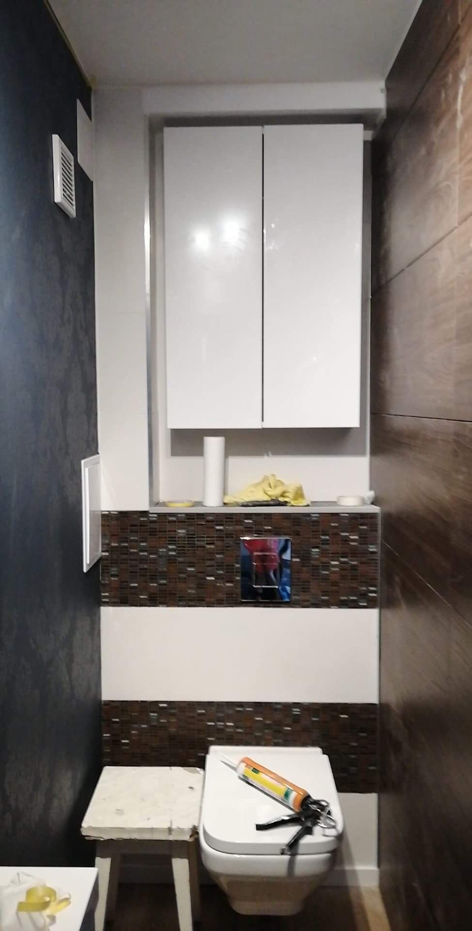 Wykończona toaleta z mozaiką, drewnopodobnymi płytkami i białymi szafkami wiszącymi; na sedesie leży pistolet do silikonu, obok stoi roboczy taboret.