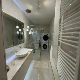 Nowoczesna łazienka z marmurowymi płytkami, złotymi akcentami, umywalką nablatową, szklaną kabiną prysznicową i pralką z suszarką w tle.