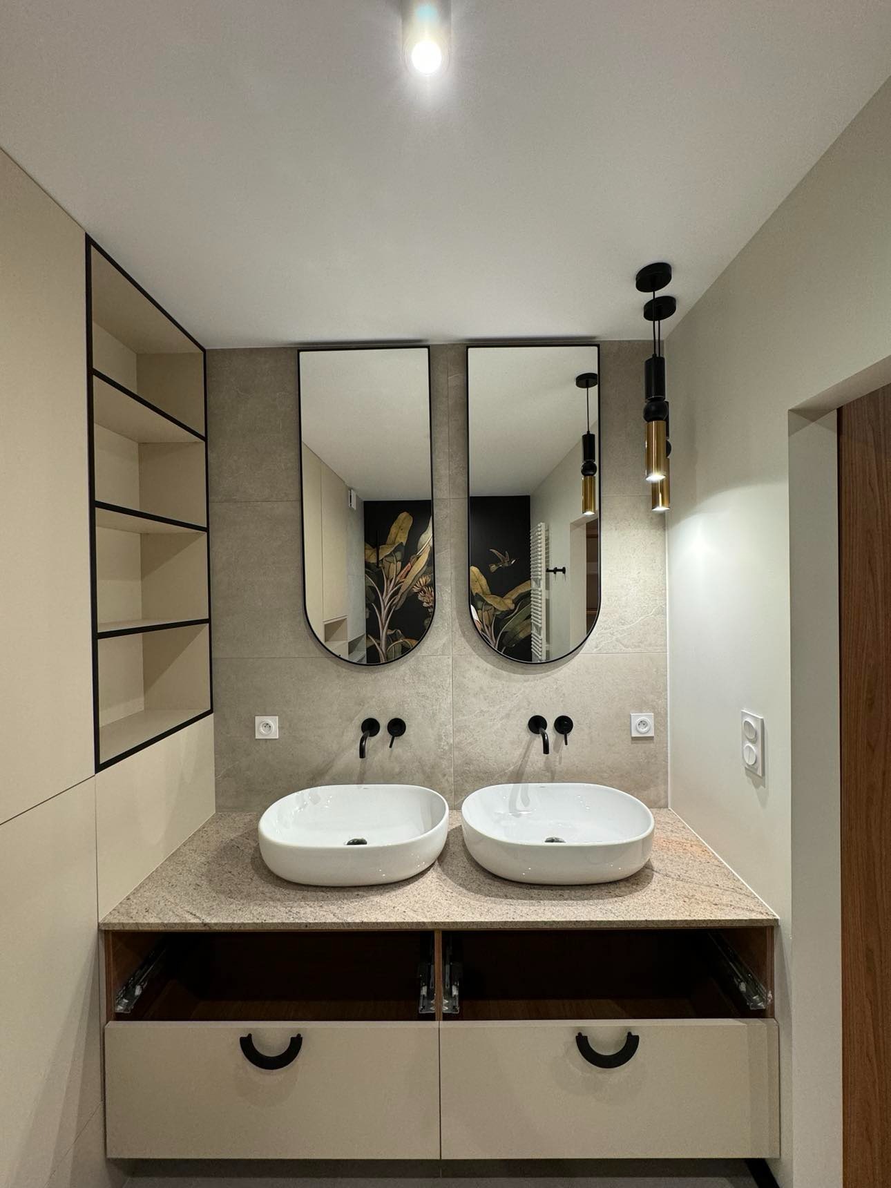 Nowoczesna łazienka z dwiema owalnymi umywalkami nablatowymi, czarnymi bateriami i lustrami w czarnych ramach, podświetlona wiszącymi lampami w kolorze czarno-złotym. W tle widoczna tapeta...