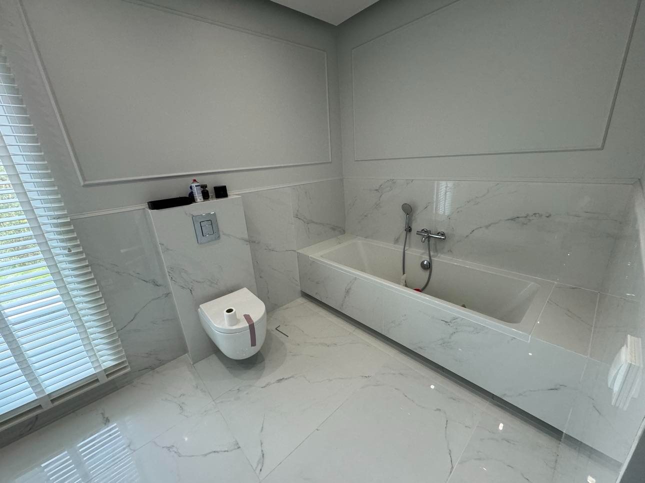 Nowoczesna łazienka wykończona białym marmurem z minimalistyczną toaletą podtynkową i wanną z hydromasażem, oświetlona światłem dziennym z okna z żaluzjami.