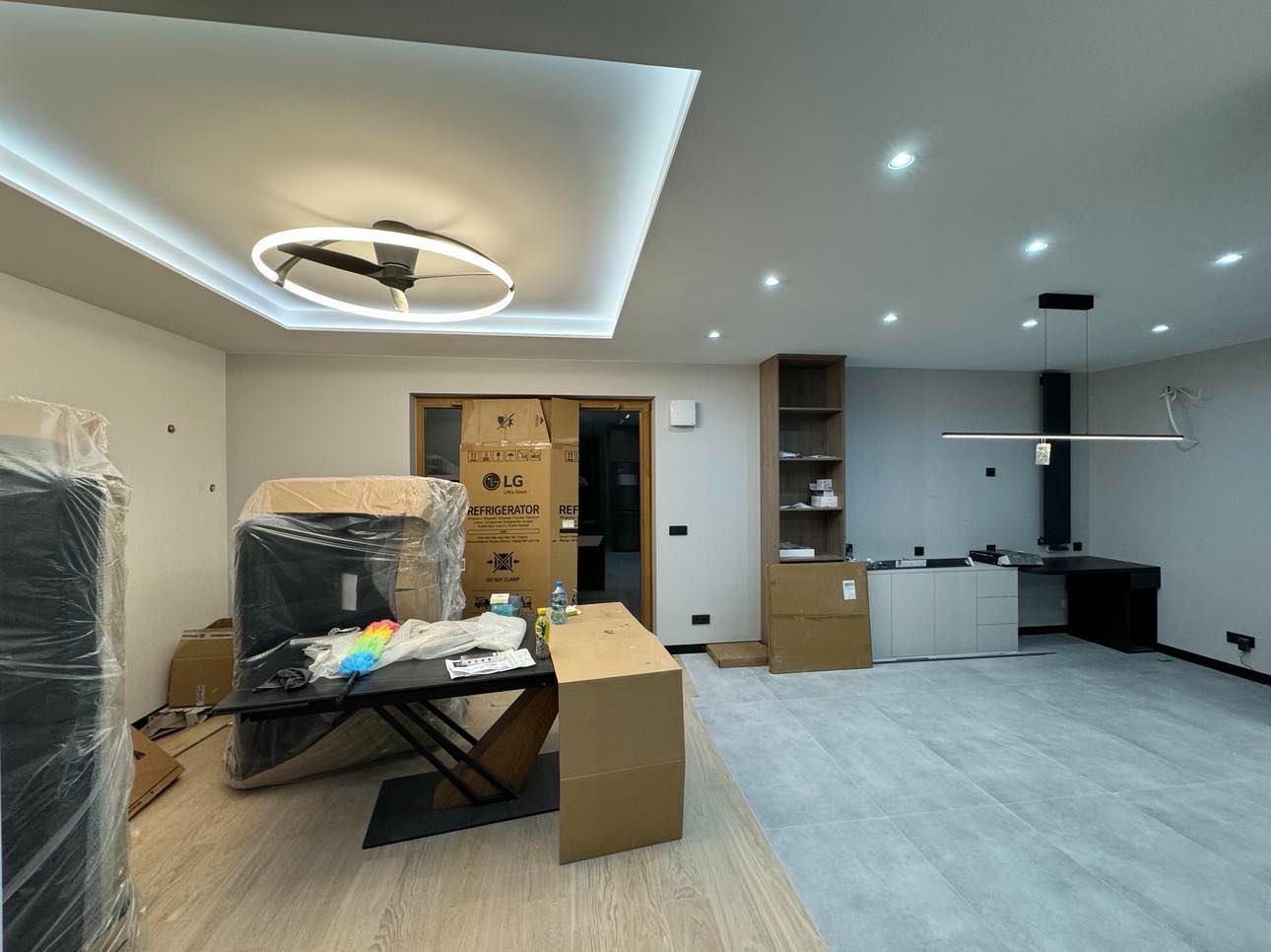 Nowoczesne wnętrze w trakcie wykańczania: podłoga z szarych płytek, zabudowa kuchenna z czarnym blatem, oświetlenie sufitowe LED, kartony z nowymi sprzętami AGD, w tym lodówka LG w folii ochronnej.