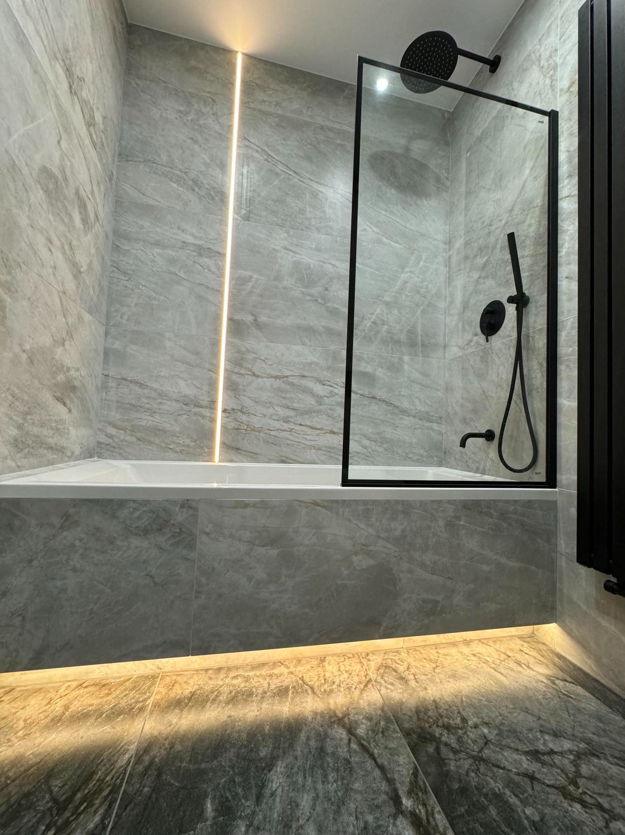 Wykończona łazienka z szarymi płytkami imitującymi marmur, podświetleniem LED i czarną armaturą. Kabina prysznicowa typu walk-in z czarną ramą.