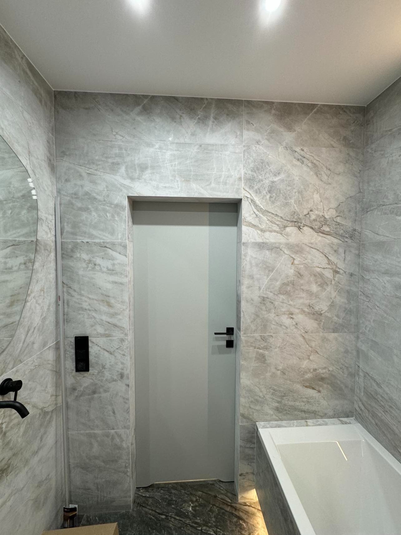 Elegancka łazienka z szarymi płytkami imitującymi marmur, czarną armaturą i prostą, szarą stolarką drzwiową. Minimalistyczny design z akcentem na naturalny kamień.