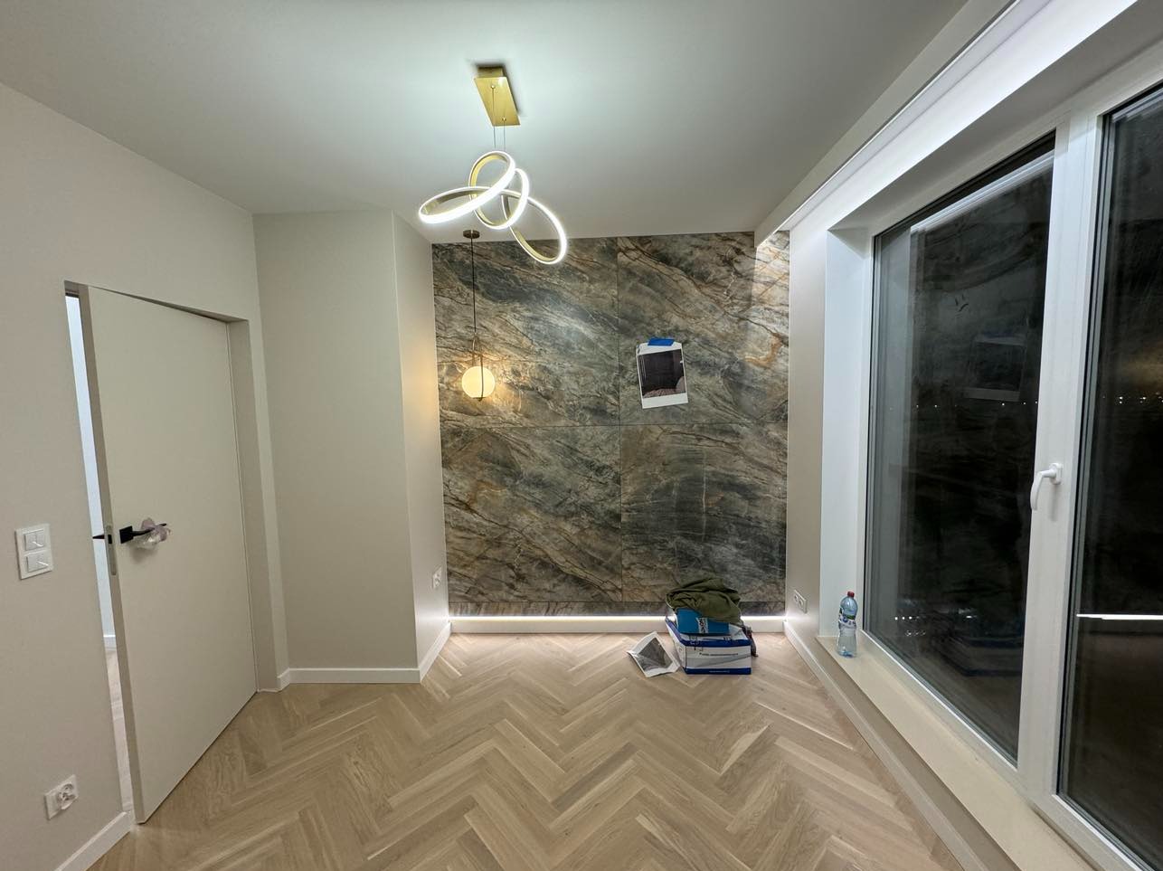 Nowoczesne wnętrze z parkietem w jodełkę i ścianą wyłożoną płytami imitującymi marmur, oświetloną listwą LED. Widoczne okno i designerska lampa sufitowa.