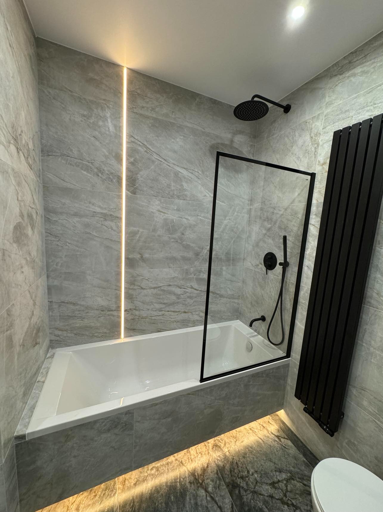 Nowoczesna łazienka z szarymi płytkami imitującymi marmur, podświetlana listwa LED i czarna armatura. Minimalistyczny design z wanną i grzejnikiem.