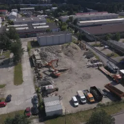 Widok z lotu ptaka na plac budowy z koparką pracującą przy hałdzie ziemi, betonowe elementy składowane obok, a w tle zabudowania przemysłowe i pojazdy budowlane.